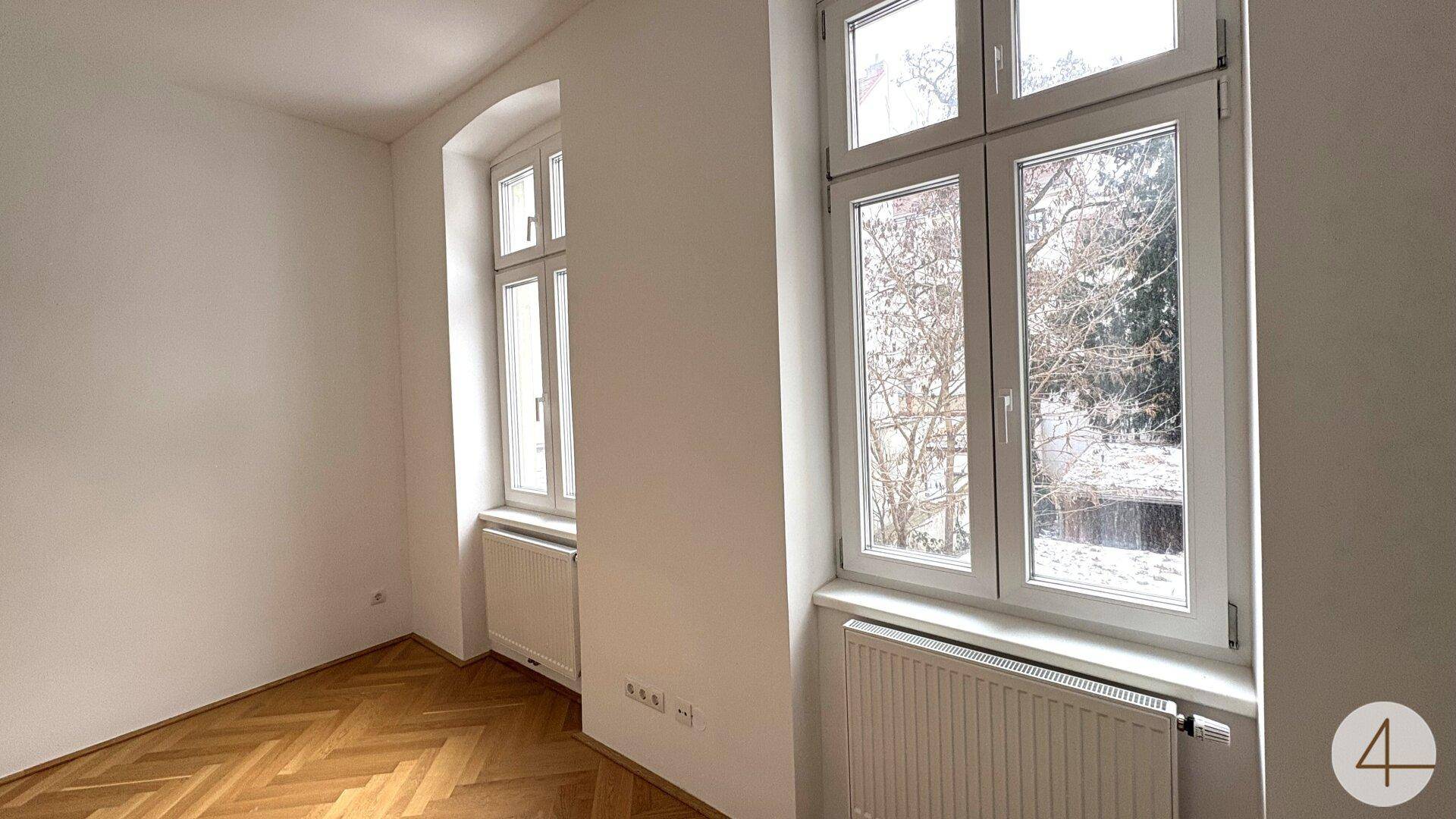 Helles Schlafzimmer mit Fischgrätparkett und Blick ins Grüne durch die großen Fenster.