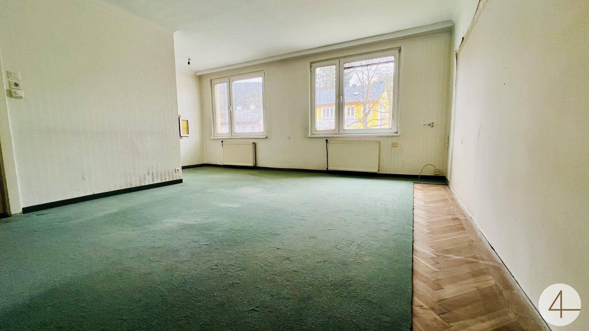 Helles Wohnzimmer mit grünem Teppichboden und Parkett, großen Fenstern und Blick ins Grüne.