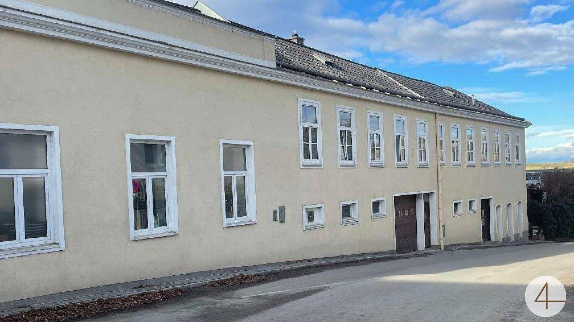 Lange, beige Gebäudefassade mit vielen Fenstern und einem Eingangsbereich an einer leicht abfallenden Straße.