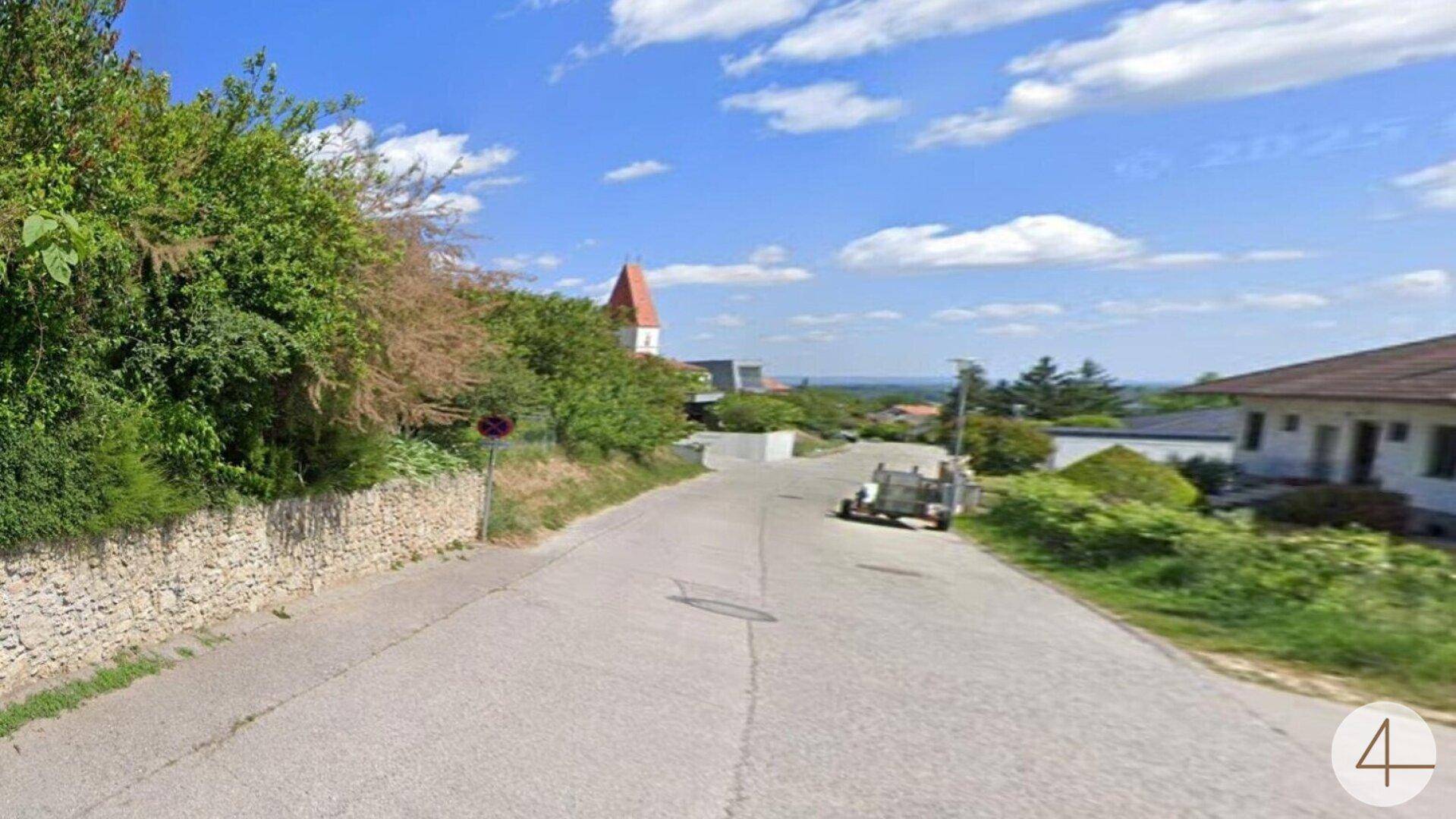 Blick auf eine ländliche Straße mit Häusern und einem Kirchturm in der Ferne unter blauem Himmel.