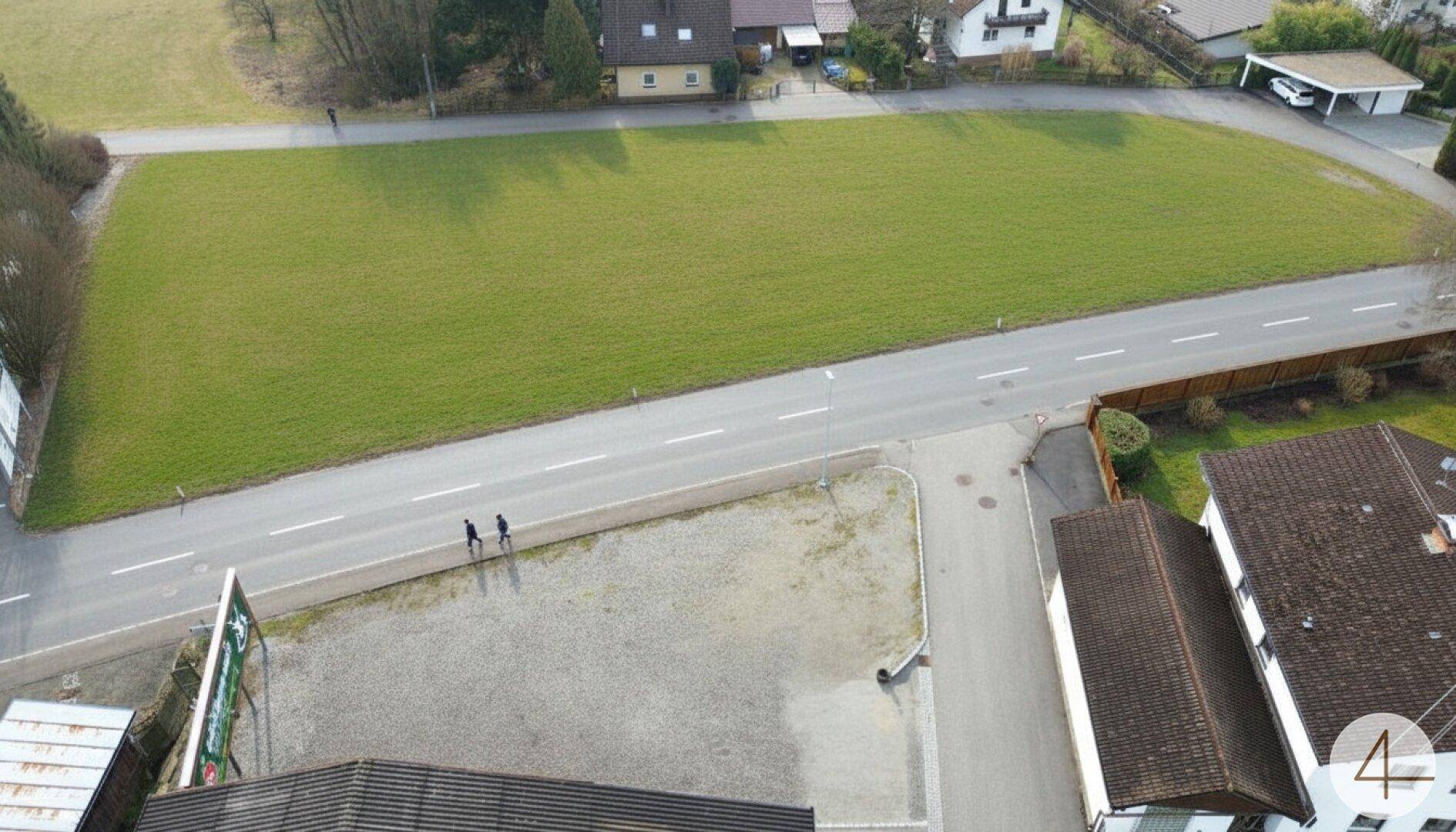 Vogelperspektive auf ein langes grünes Grundstück entlang einer Straße mit Fußgängern und Gebäuden.