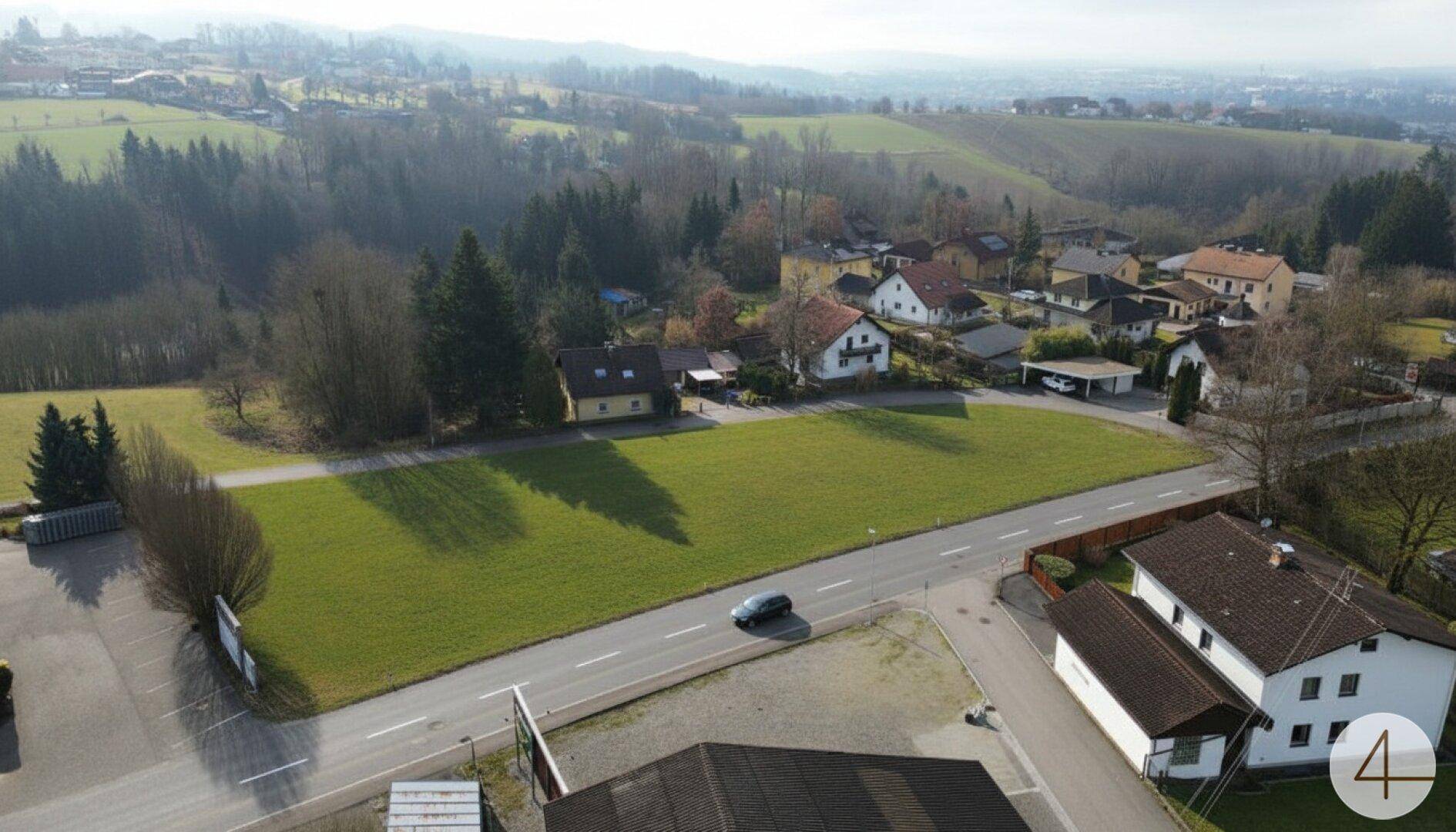 Weitläufige Luftaufnahme eines grünen Grundstücks mit Straße und hügeliger Landschaft im Hintergrund.