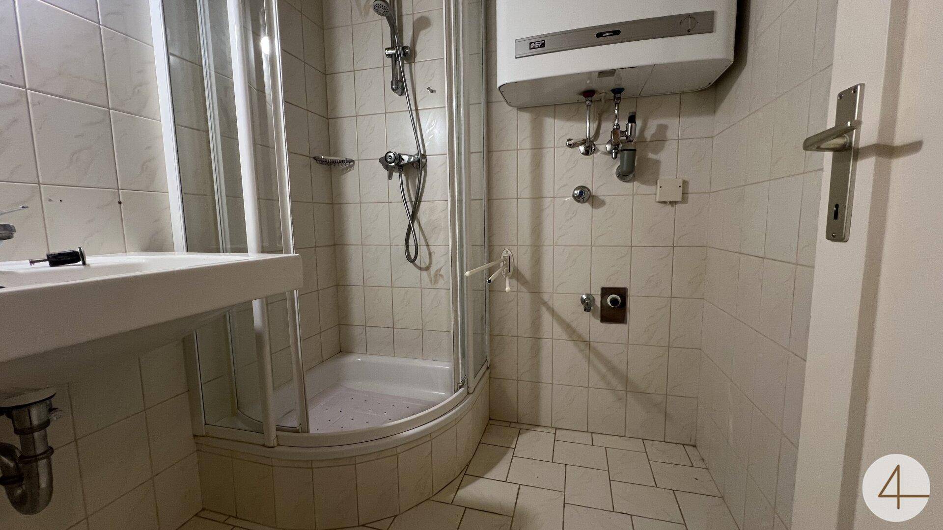 Kleines Badezimmer mit Duschkabine, Waschbecken und Warmwasserbereiter, benötigt Modernisierung.