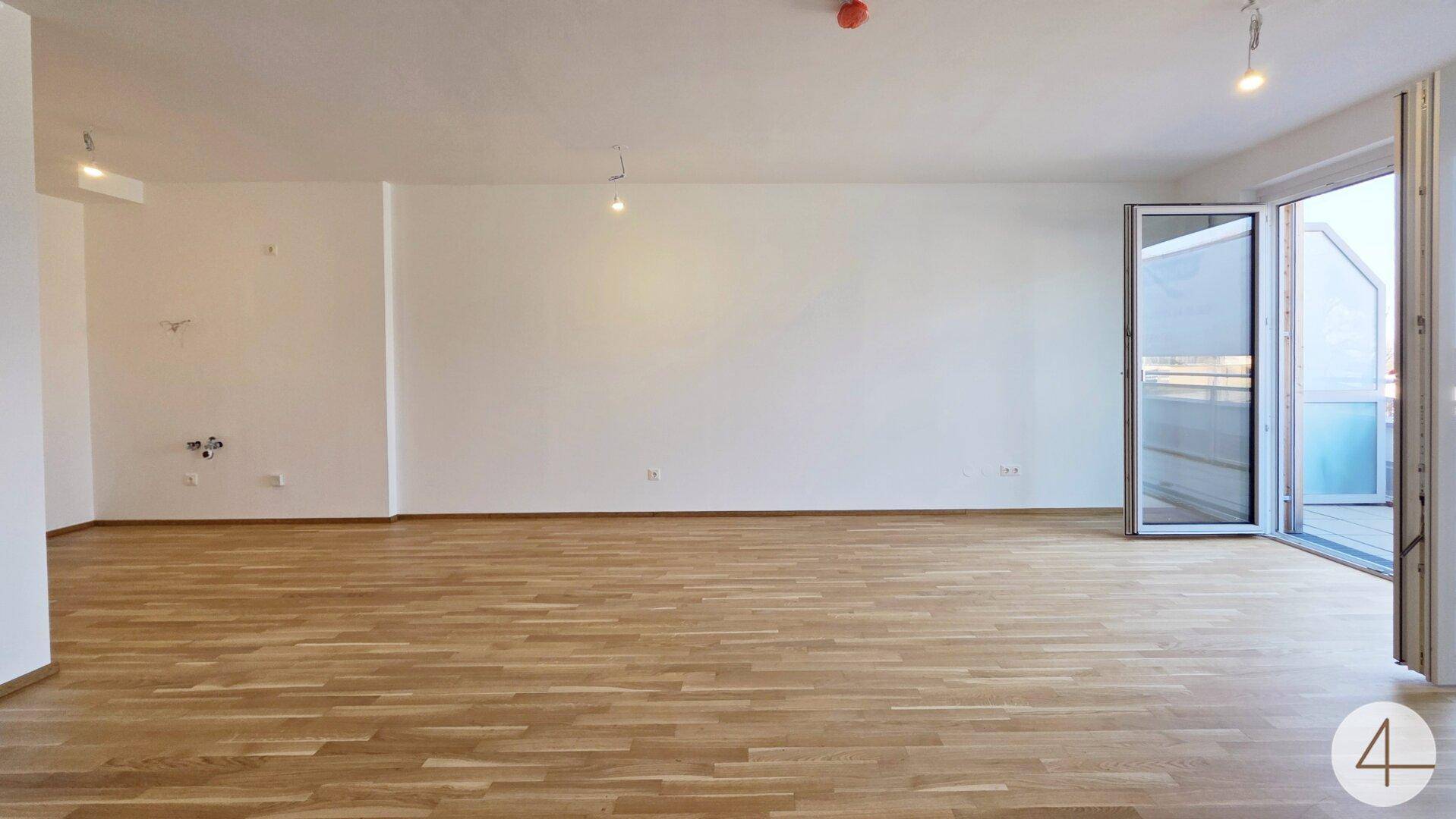 Leeres Wohnzimmer mit hellem Parkettboden und breiter Fensterfront zum Außenbereich.