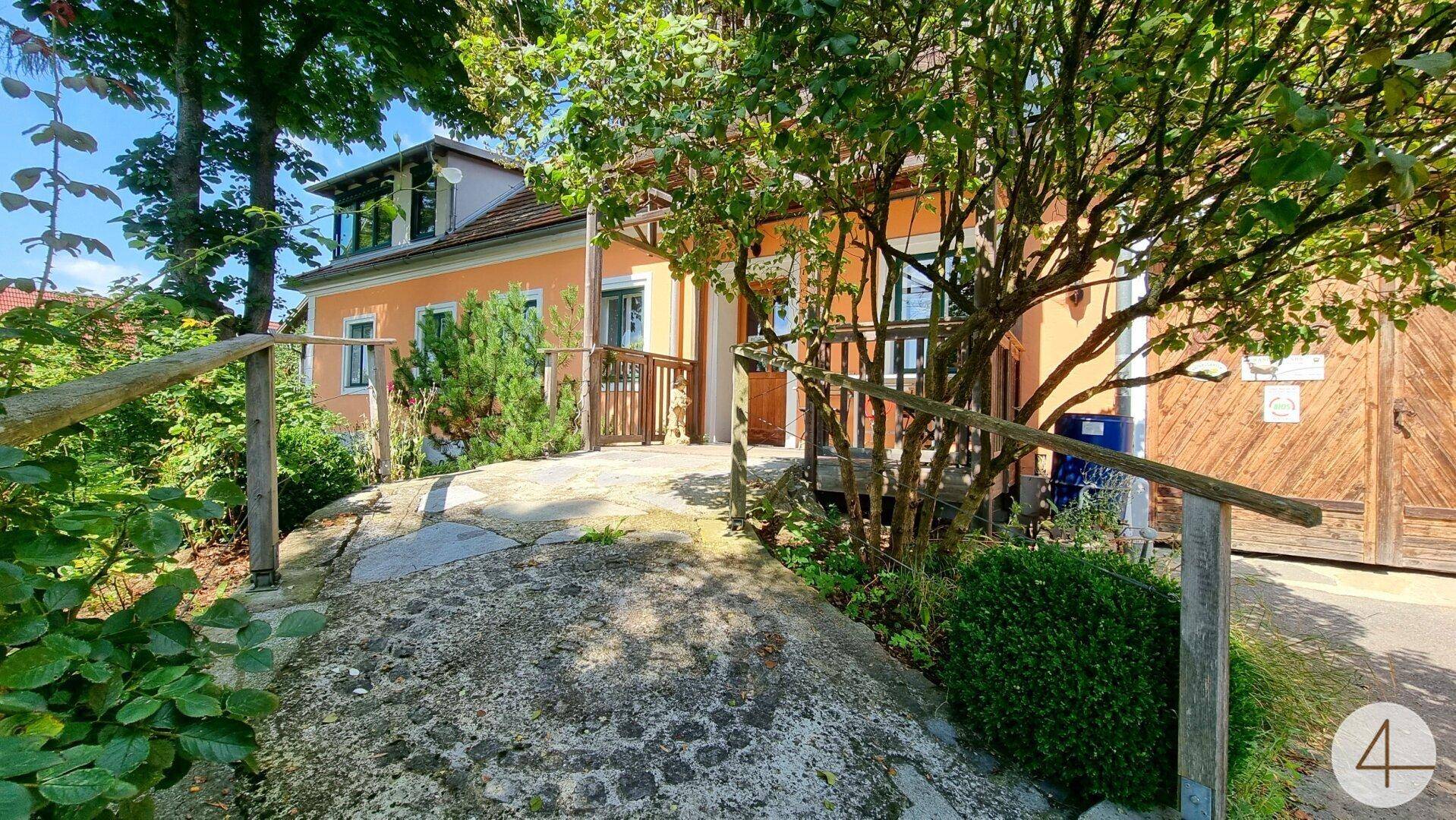 Charmantes orangefarbenes Haus mit Holzbalkon, umgeben von üppigem Grün und einem kleinen Weg.