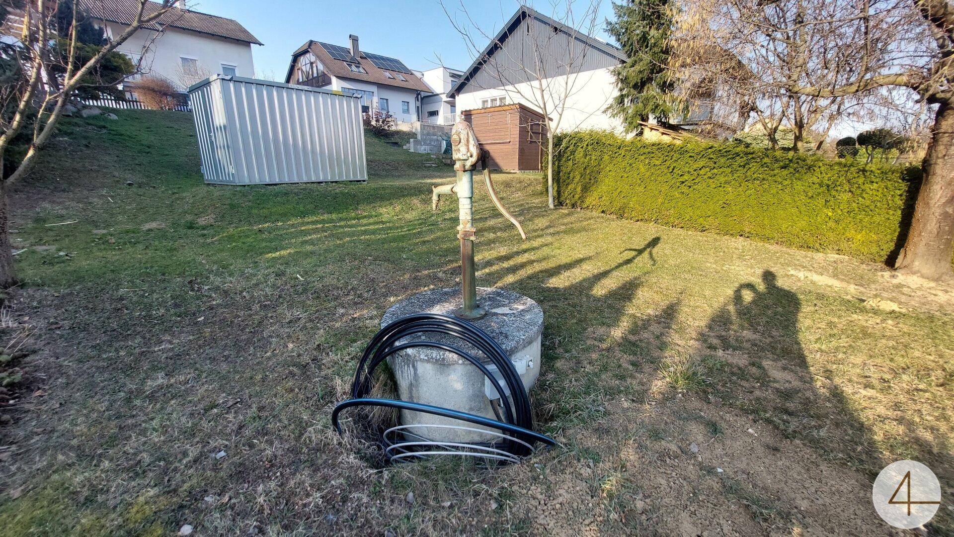 Gartenbereich mit Rasen, einem alten Wasserbrunnen und einem Metallschuppen im Hintergrund.
