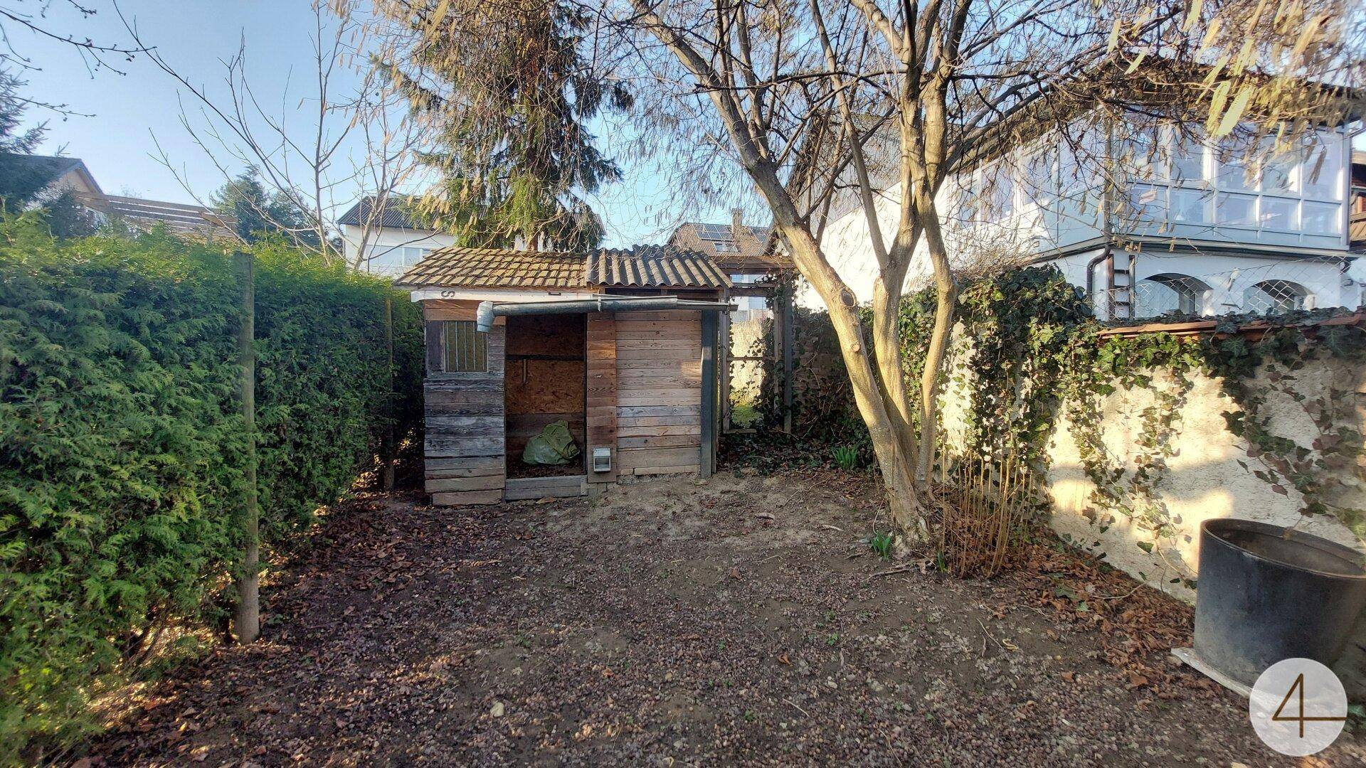 Rustikaler Holzunterstand im Garten, umgeben von hohen Hecken und einer alten Steinmauer.
