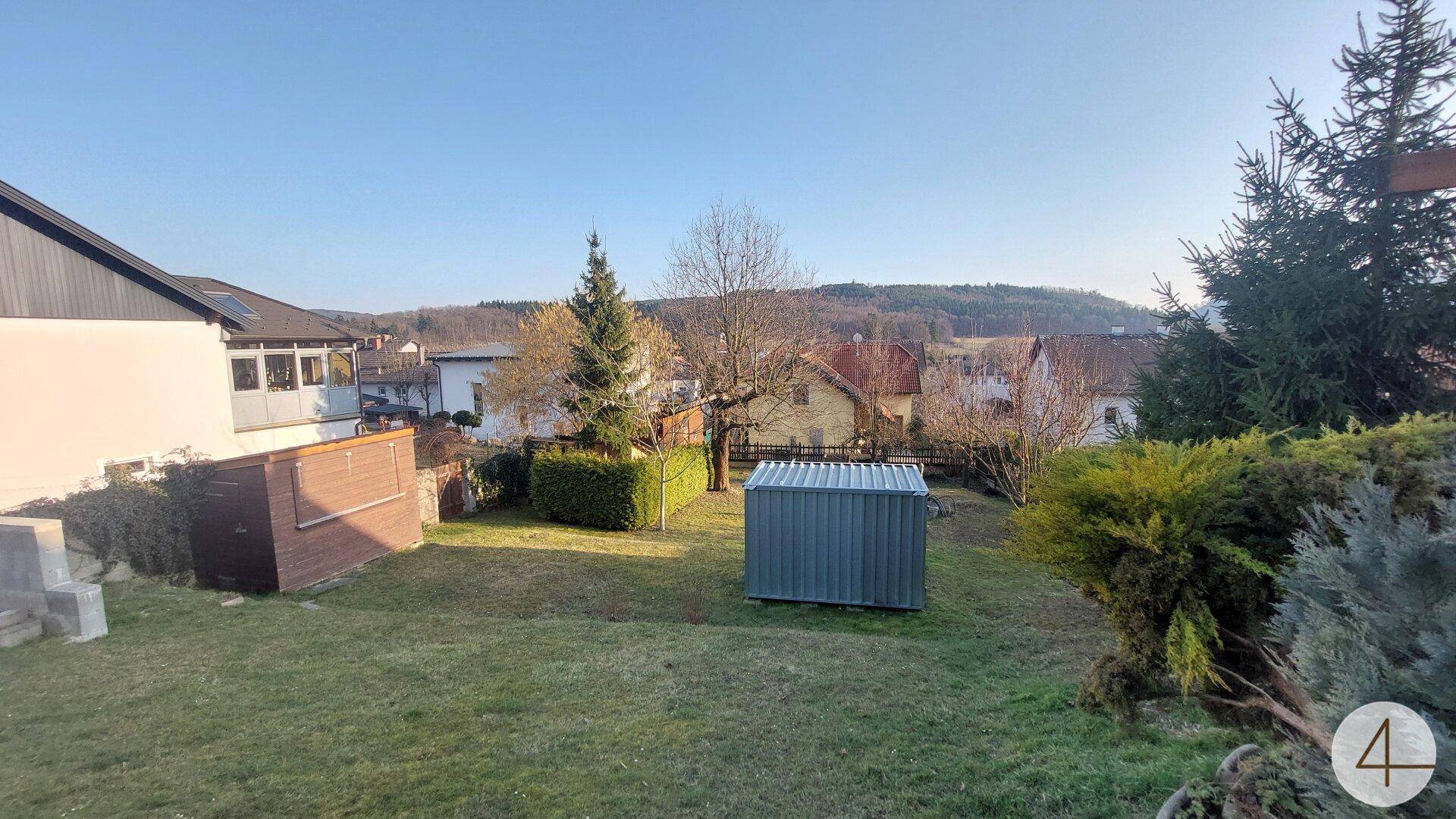 Weitläufiger Garten mit Rasen, zwei Schuppen und Panoramablick auf die grüne Umgebung und Hügel.