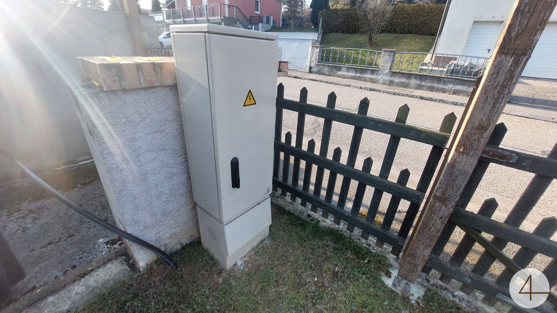 Elektrischer Verteilerkasten am Zaun neben der Straße, mit Warnzeichen versehen und sonnigem Licht.