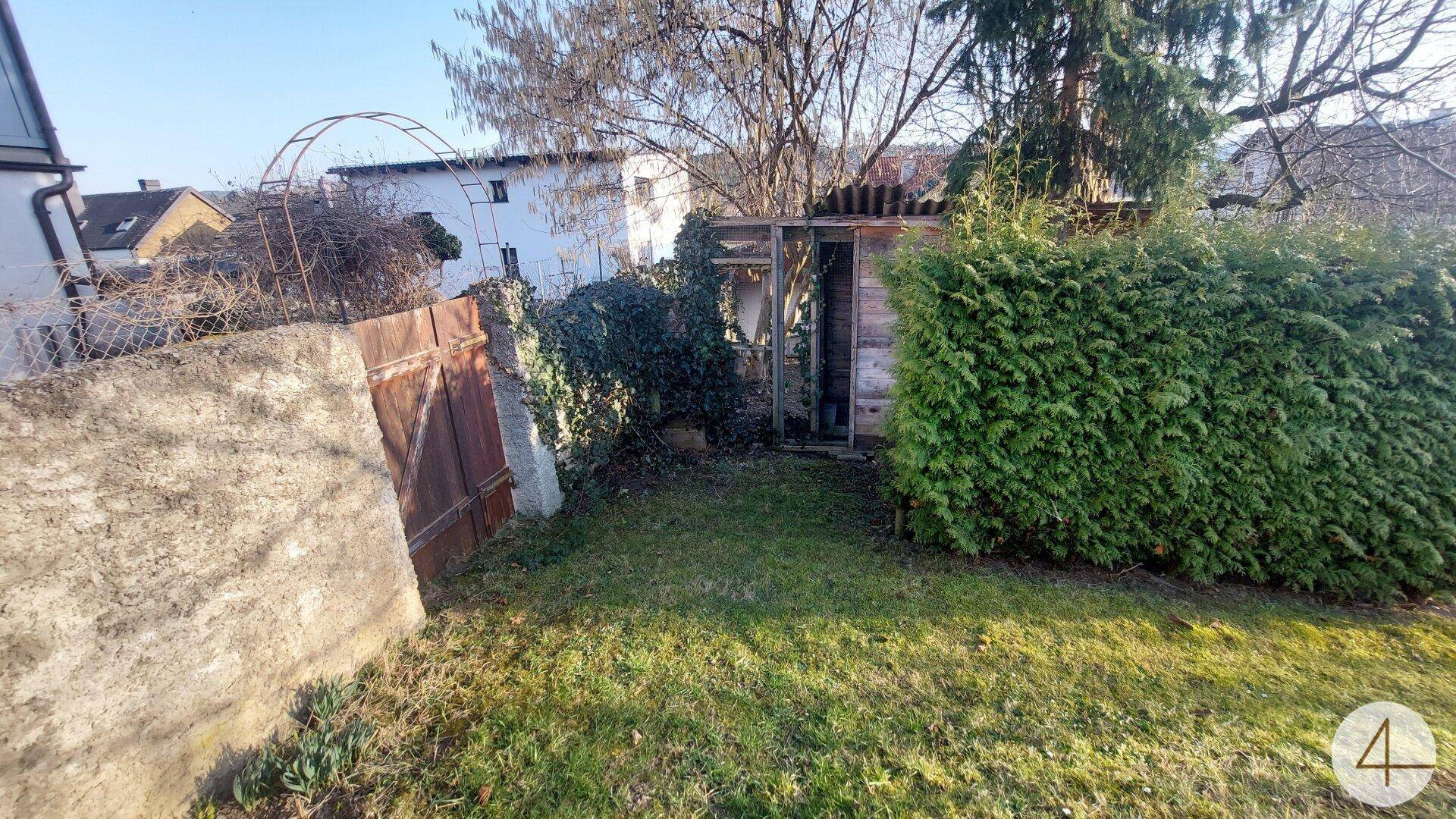 Gartenbereich mit Rasen, einem rustikalen Holzunterstand und einer alten Steinmauer mit Tor.