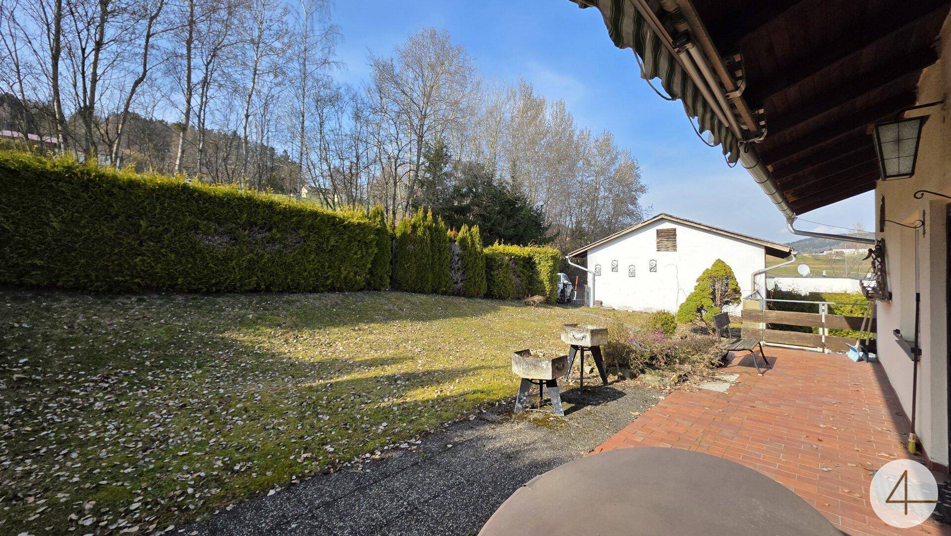 Garten mit Rasenfläche, hoher Hecke und einer gepflasterten Terrasse mit Grillbereich.
