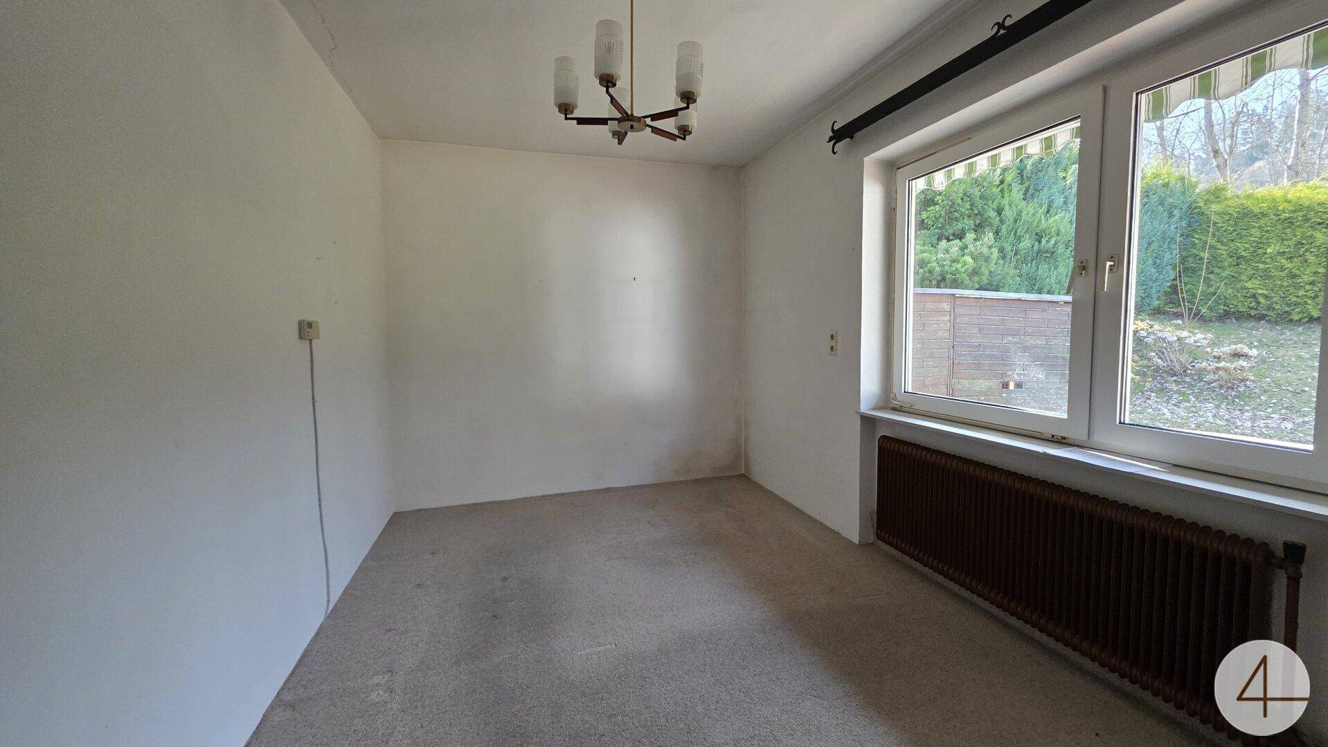 Leeres Zimmer mit großem Fenster, Heizkörper und Blick auf eine grüne Hecke.