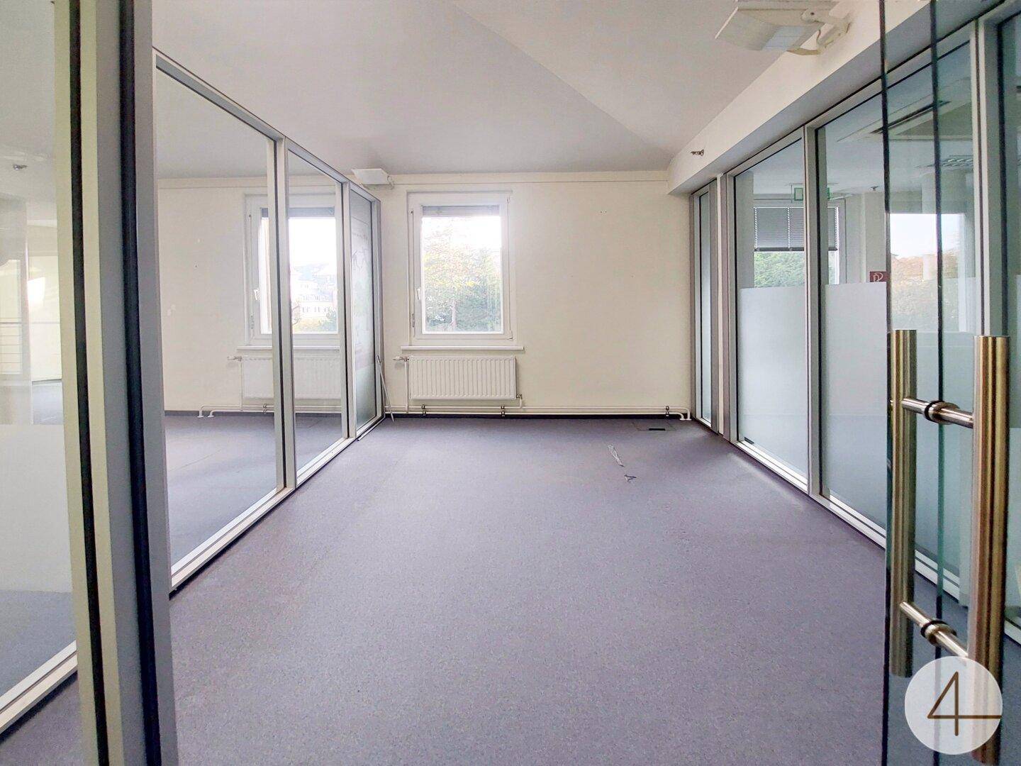 Innenansicht eines Bürozimmers mit Teppichboden und Fenster, gesehen durch Glaswände.