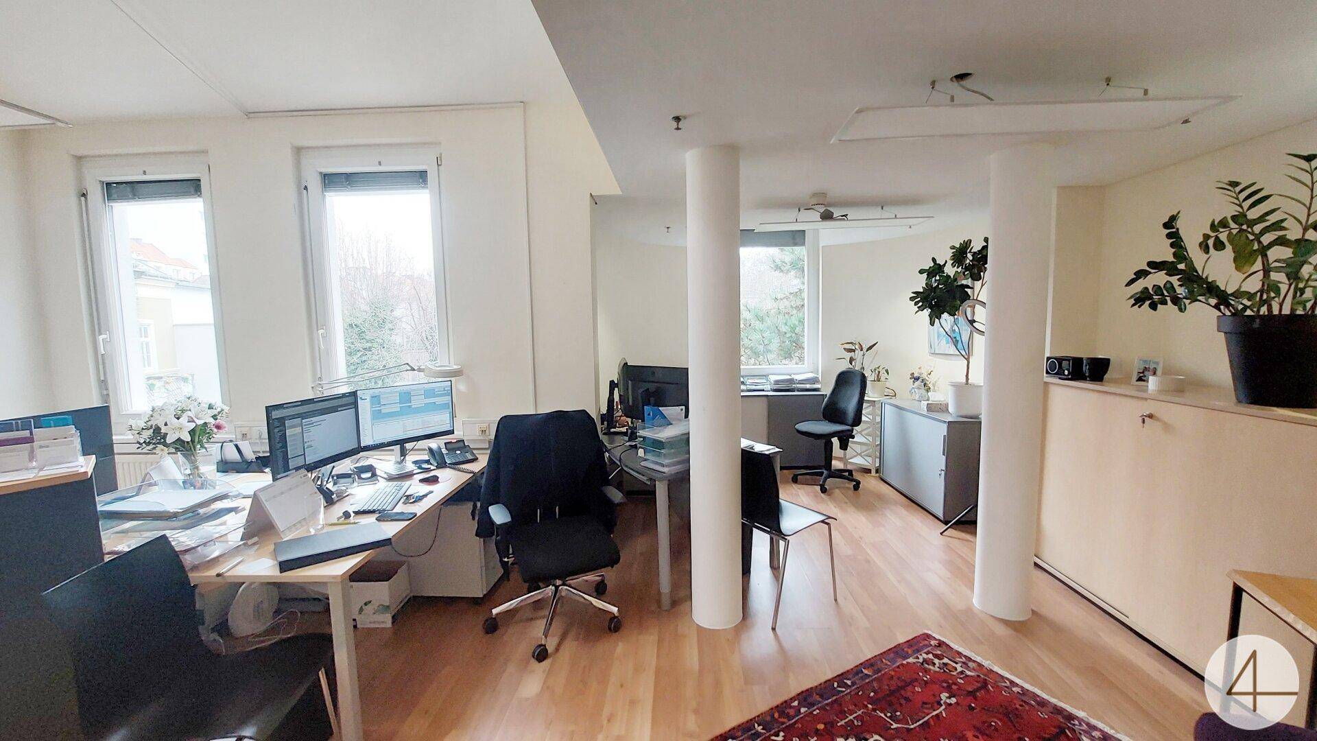 Offenes Büro mit mehreren Arbeitsplätzen, Holzboden und großen Fenstern, die viel Licht hereinlassen.