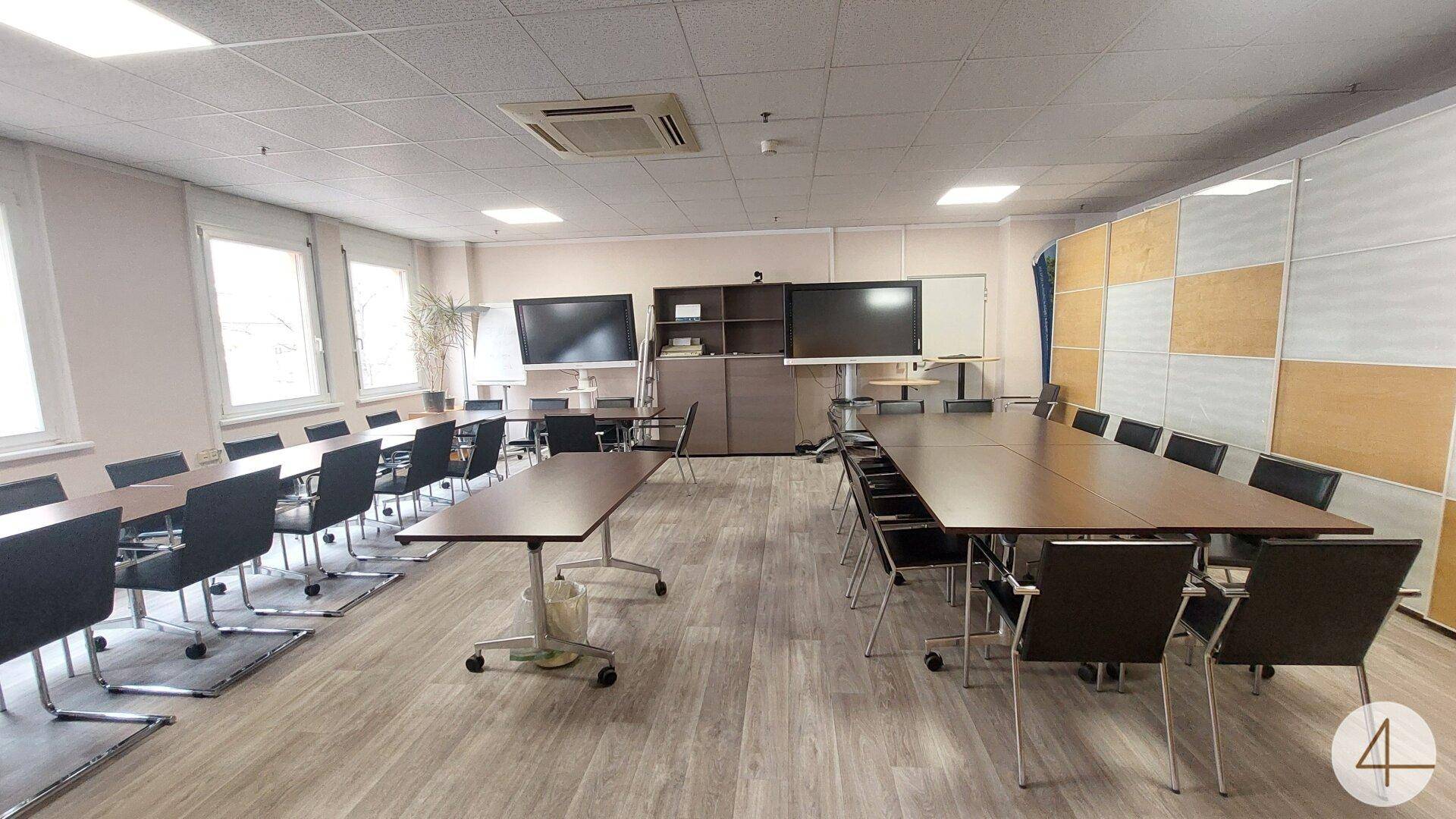 Großer Konferenzraum mit mehreren Tischen, Stühlen, Bildschirmen und einer modernen Wandverkleidung.