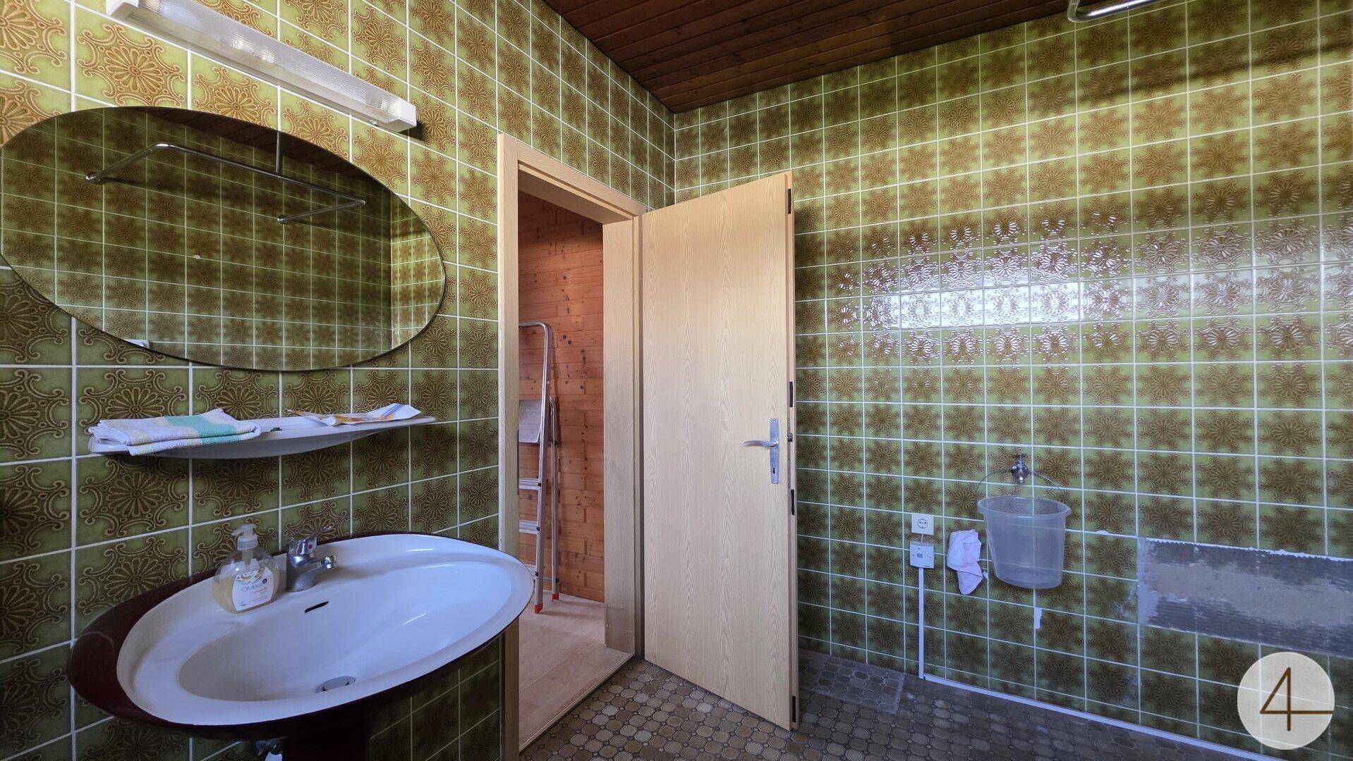 Badezimmer im Stil der 80er Jahre mit auffälligen grünen Fliesen, Waschbecken und ovalem Spiegel. Die Holzdecke verleiht dem Raum einen rustikalen Charme.