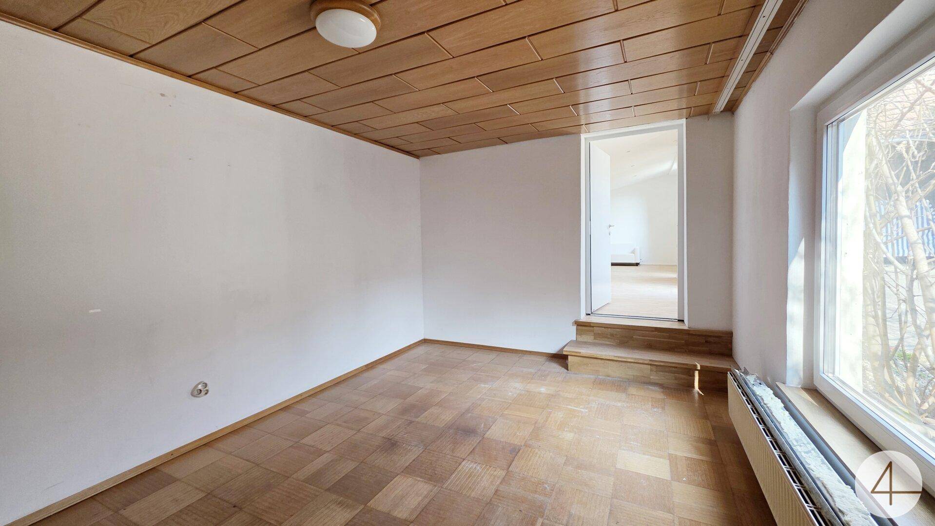Heller Raum mit Parkettboden, Holzdecke und Zugang zu einem weiteren Zimmer.