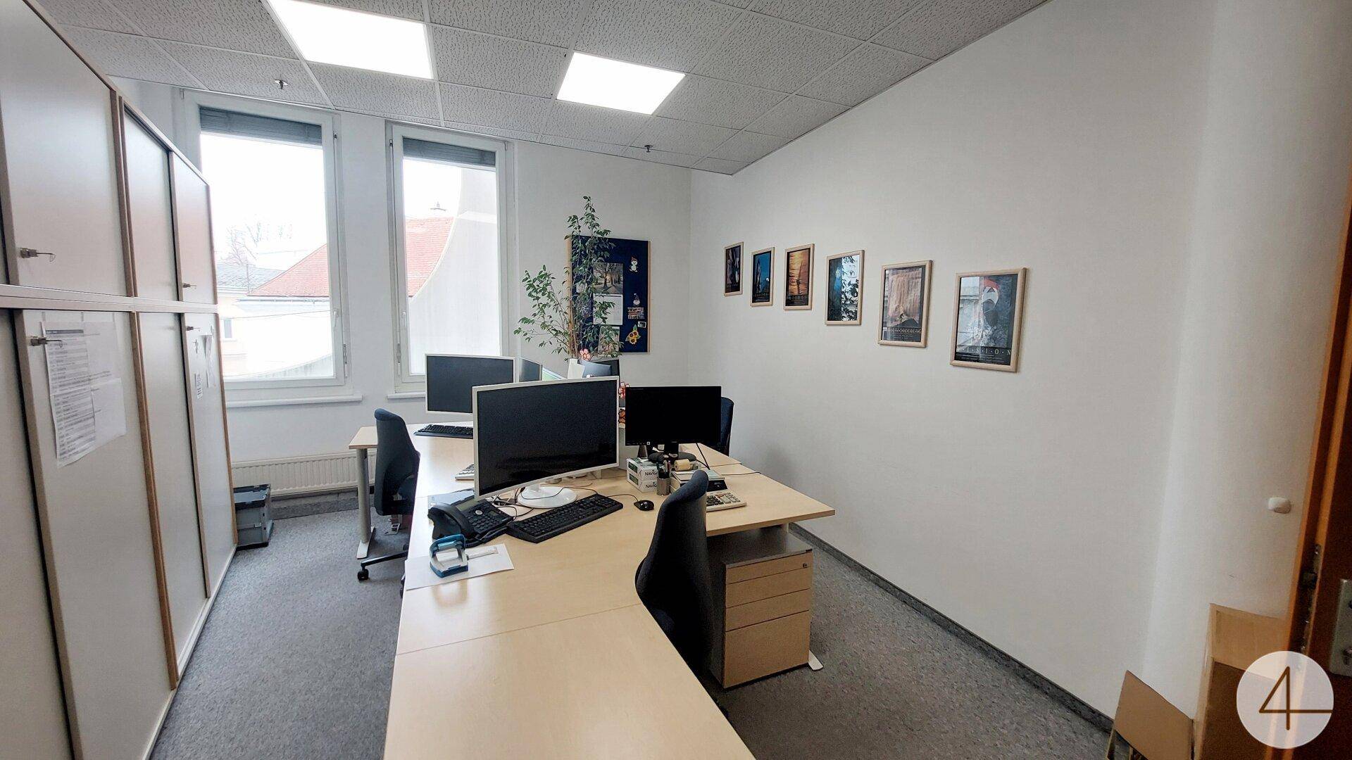 Helles Büro mit mehreren Schreibtischen, Teppichboden und Fenstern mit Blick auf die Umgebung.