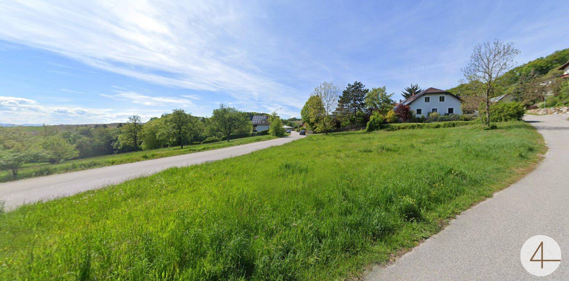 Panoramablick über ein sonniges Grundstück mit Straße und bewaldeter Landschaft am Horizont.