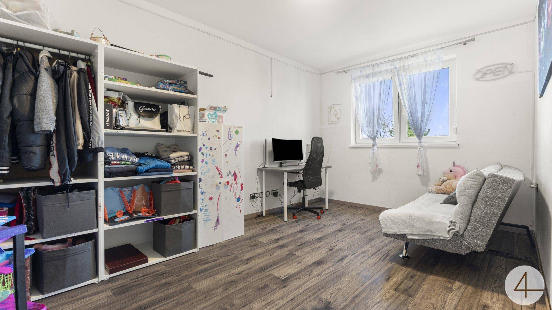 Vielseitiges Zimmer mit Schlafsofa, offenem Regal und Schreibtisch am Fenster.