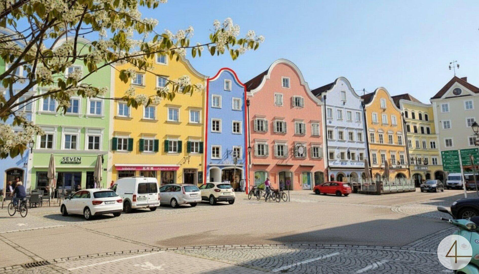 Bunte historische Gebäude an einem Marktplatz mit geparkten Autos und Fußgängern an einem sonnigen Tag.