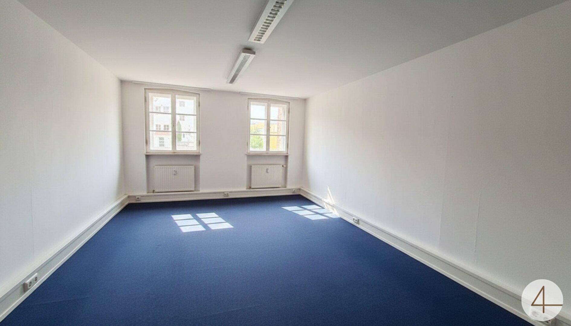Heller Büroraum mit zwei Fenstern, blauem Teppichboden und weißen Wänden, durch Sonnenlicht beleuchtet.