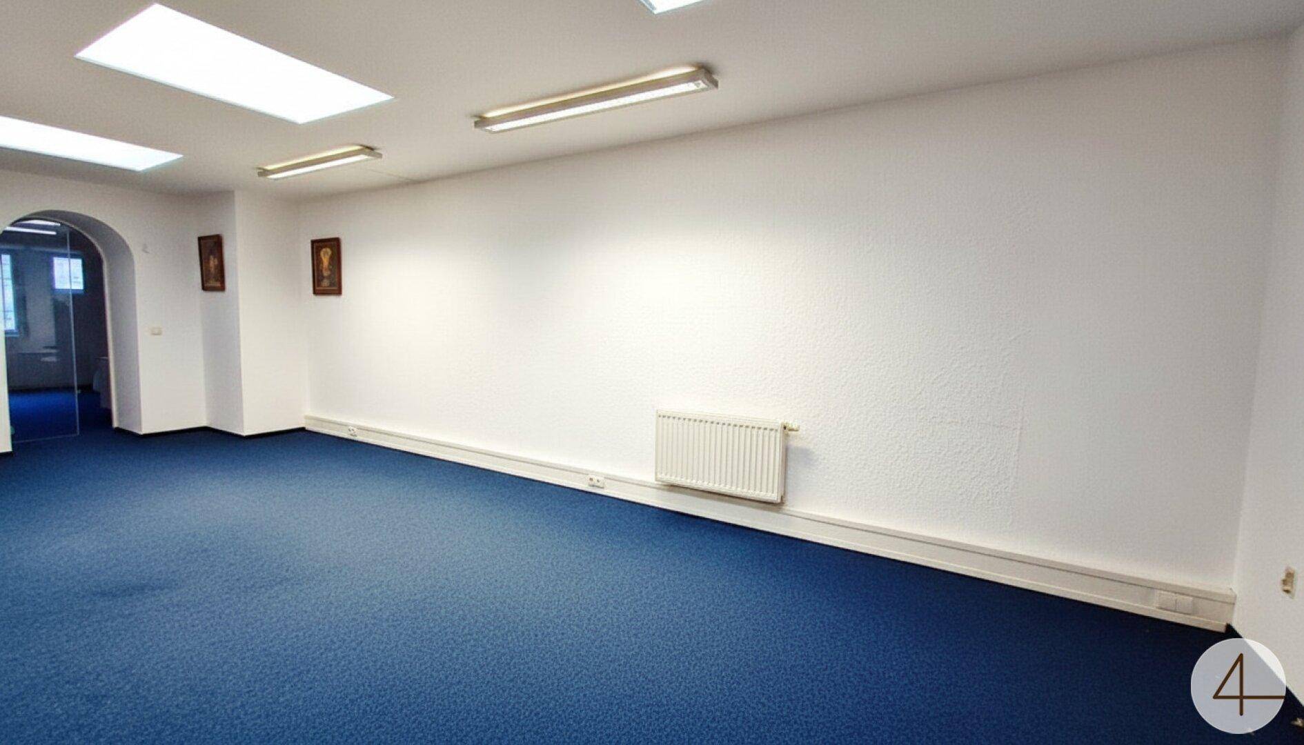 Großer, heller Büroraum mit blauem Teppichboden, weißen Wänden und modernen Deckenleuchten.