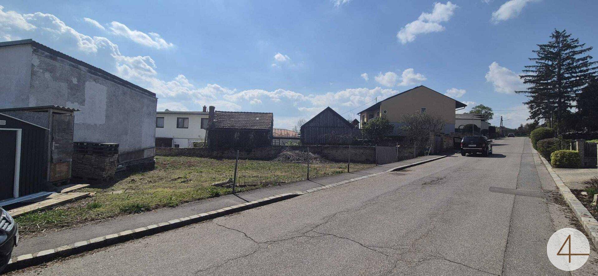 Ländliche Straße mit Bürgersteig und unbebautem Grundstück, umgeben von Wohnhäusern.