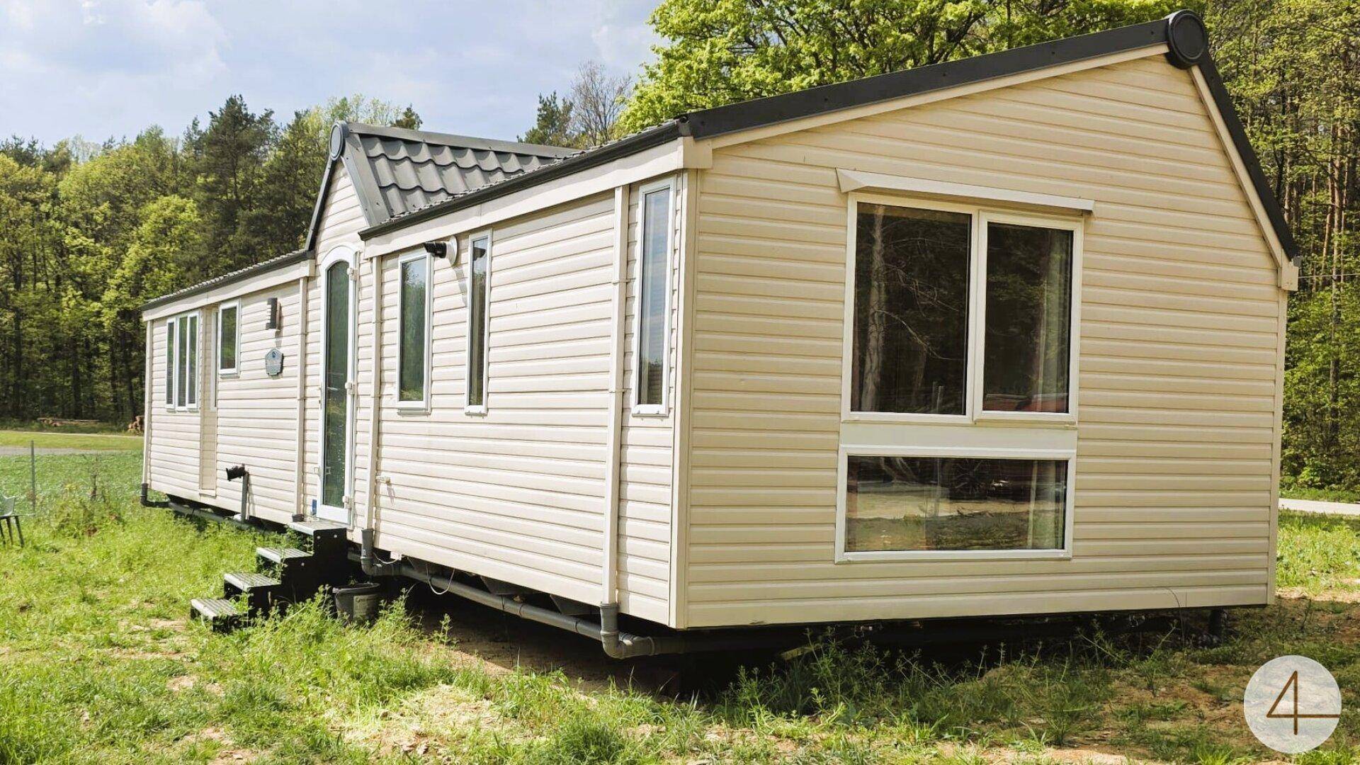 Freistehendes Mobilheim mit beiger Fassade und dunklem Dach, gelegen auf einer grünen Wiese am Waldrand.