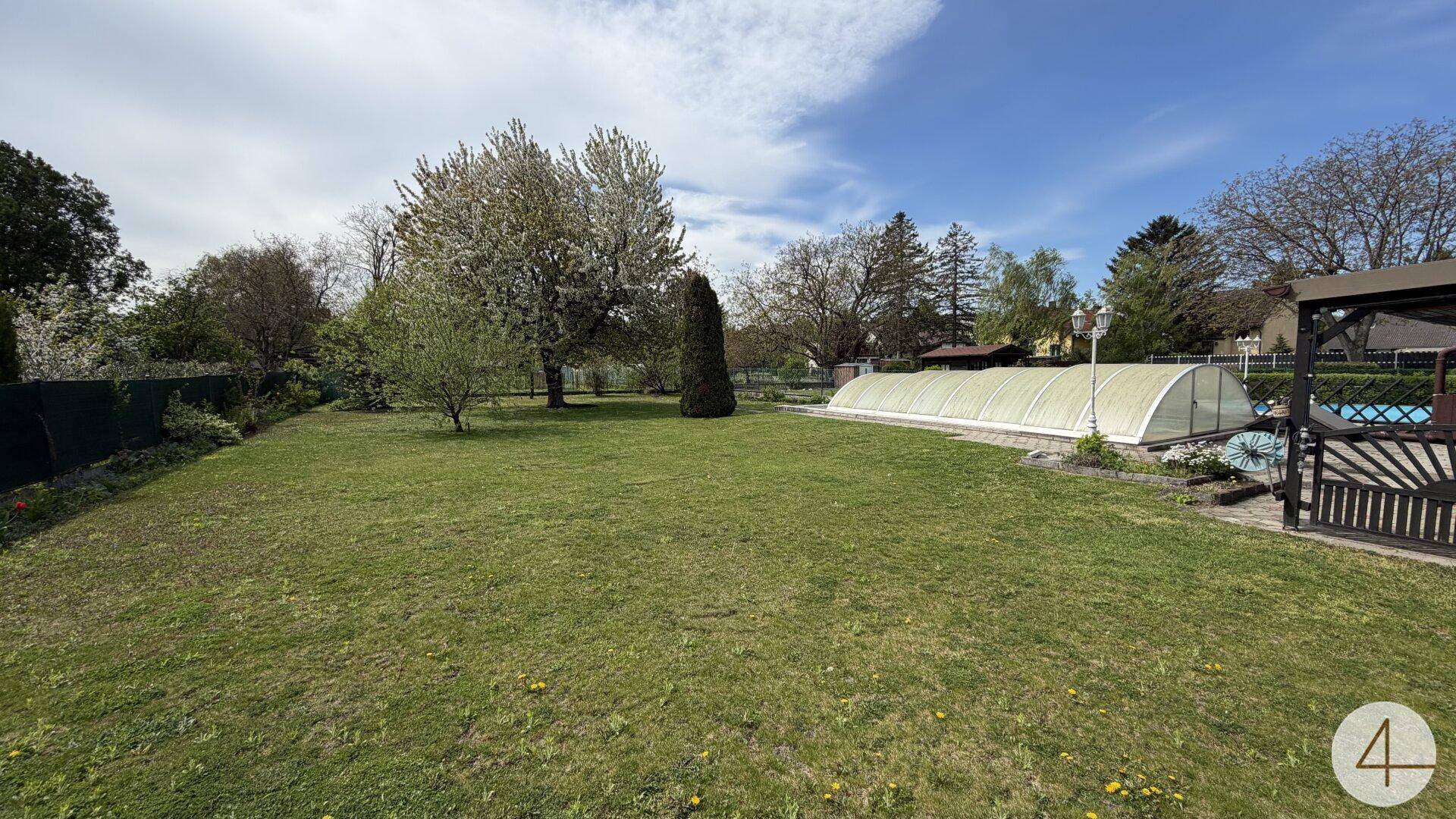 Weitläufiger Garten mit blühenden Bäumen, großer Rasenfläche und einem überdachten Swimmingpool.