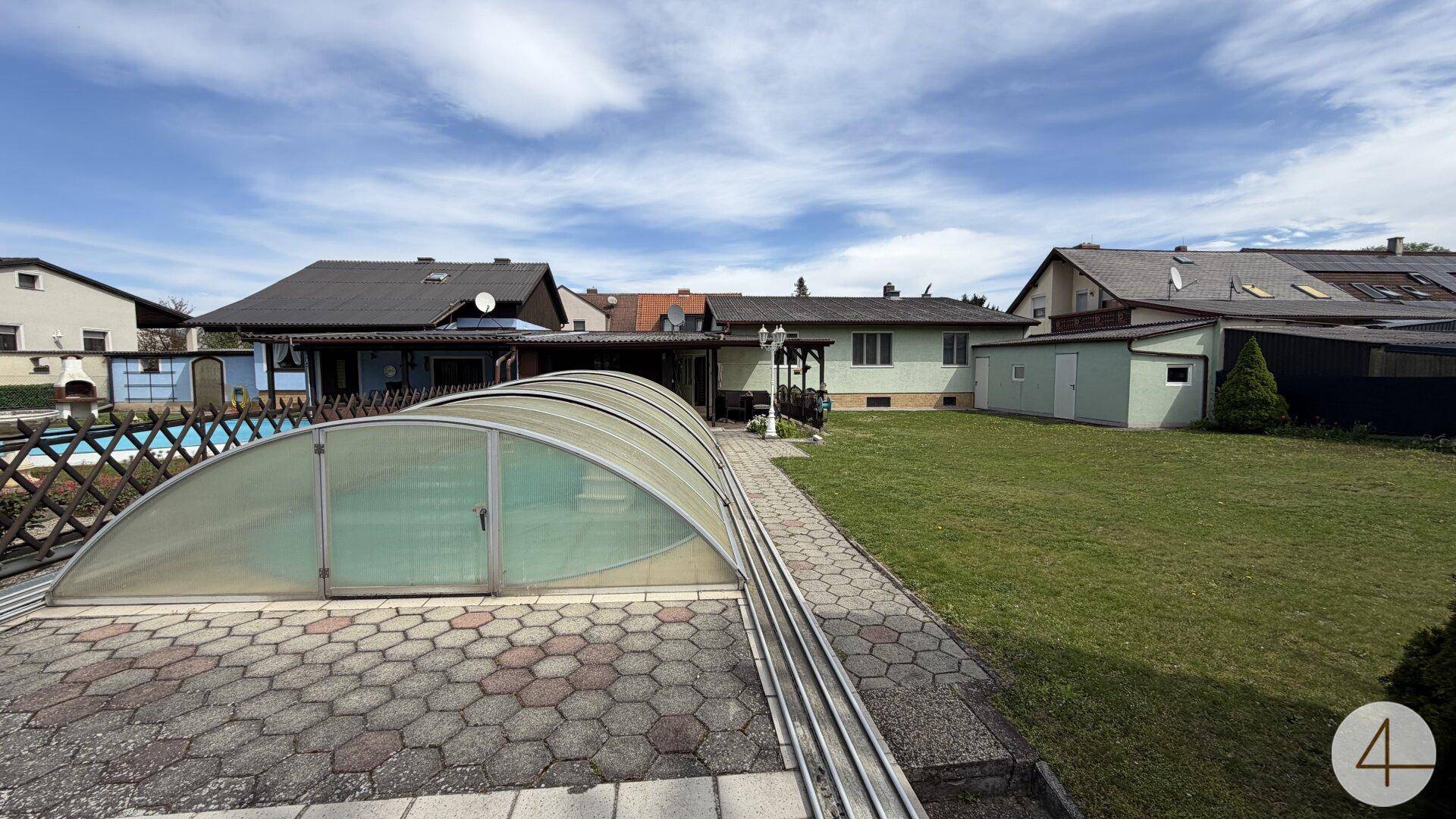 Gepflasterter Bereich um den überdachten Pool mit angrenzender Rasenfläche und Blick auf das Haus.