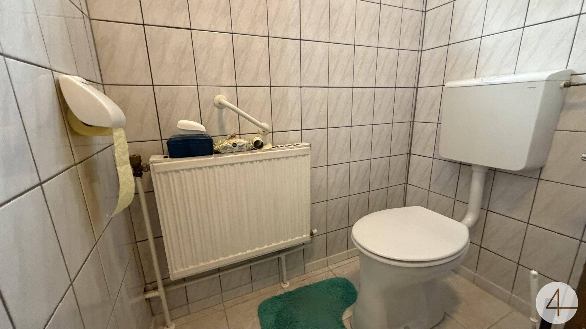 Kleines WC mit hellen Fliesen, Toilette und Heizkörper, ausgestattet mit Haltegriffen.