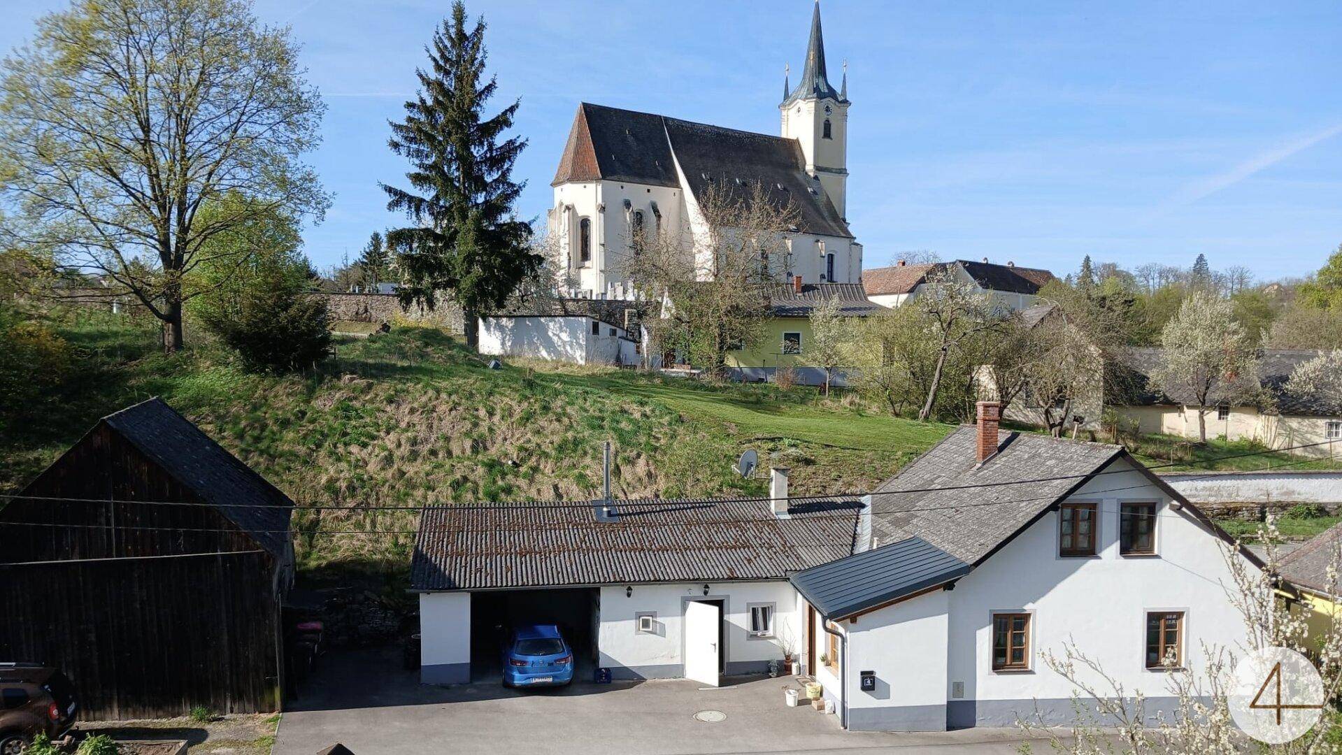 Blick auf ein traditionelles Wohnhaus mit Garage und Garten, im Hintergrund eine historische Kirche in ländlicher Umgebung.