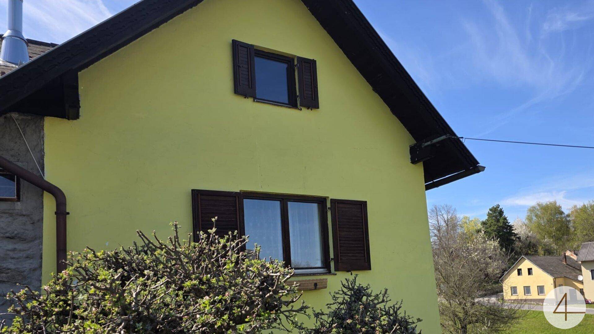Seitenansicht eines Hauses mit gelber Fassade, braunen Fensterläden und Satteldach unter blauem Himmel.
