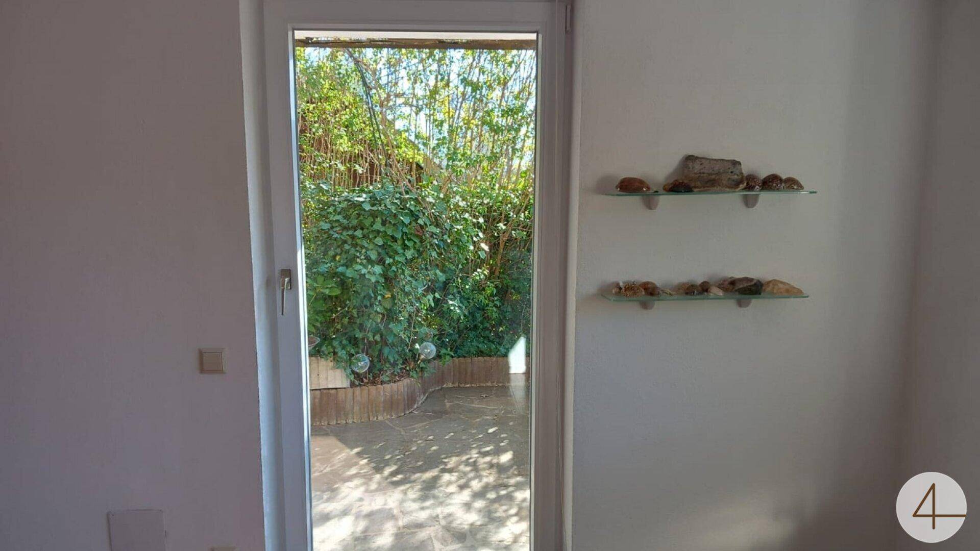 Heller Innenraum mit Zugang zu einer kleinen, begrünten Terrasse und dekorativen Glasregalen an der Wand.
