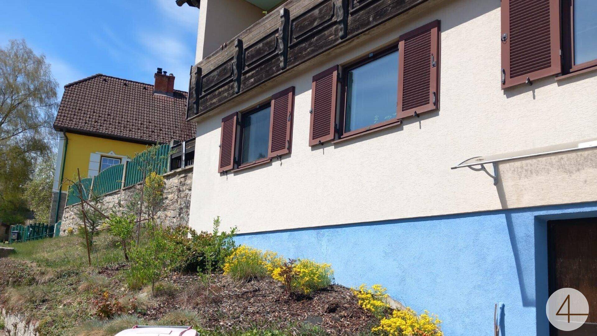 Detailansicht der Hausfassade mit braunen Fensterläden, blauem Sockel und kleinen Sträuchern im Vorgarten.