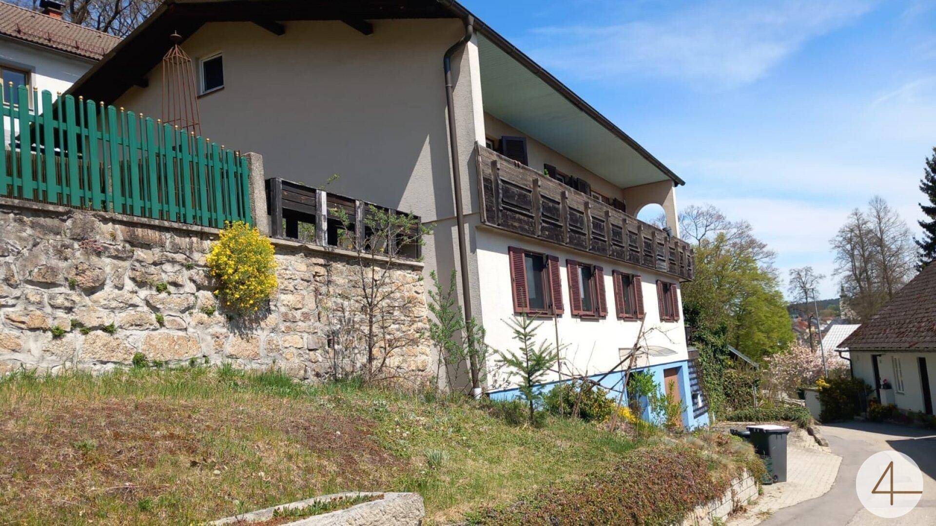 Hausfassade an einem Hang mit Steinmauer, grünem Zaun und einem Holzbalkon im oberen Bereich.