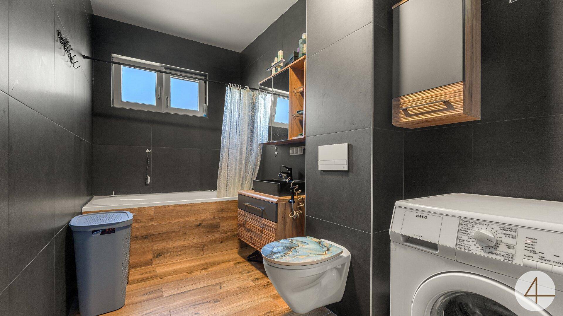 Modernes Badezimmer mit dunklen Fliesen, Holzoptikboden, Badewanne und Waschmaschine.