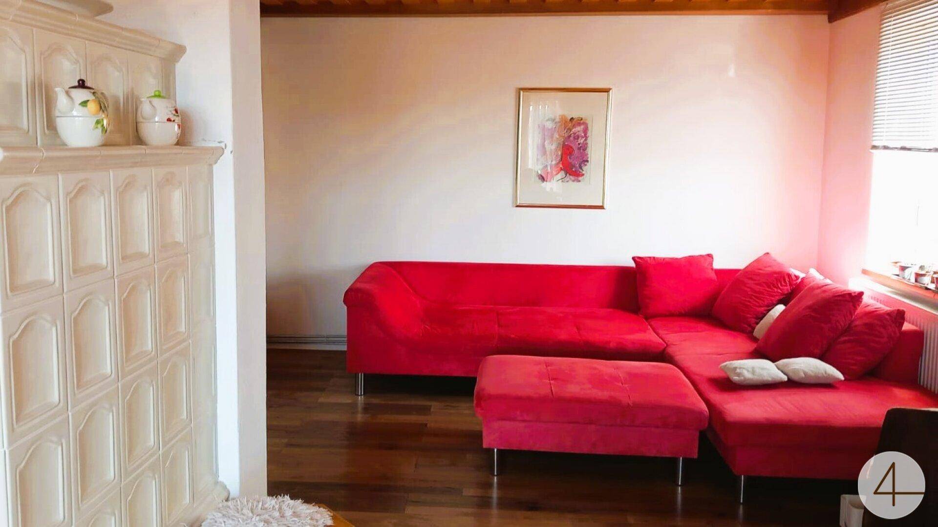 Gemütlicher Wohnbereich mit großem rotem Ecksofa und traditionellem Kachelofen an der Wand.