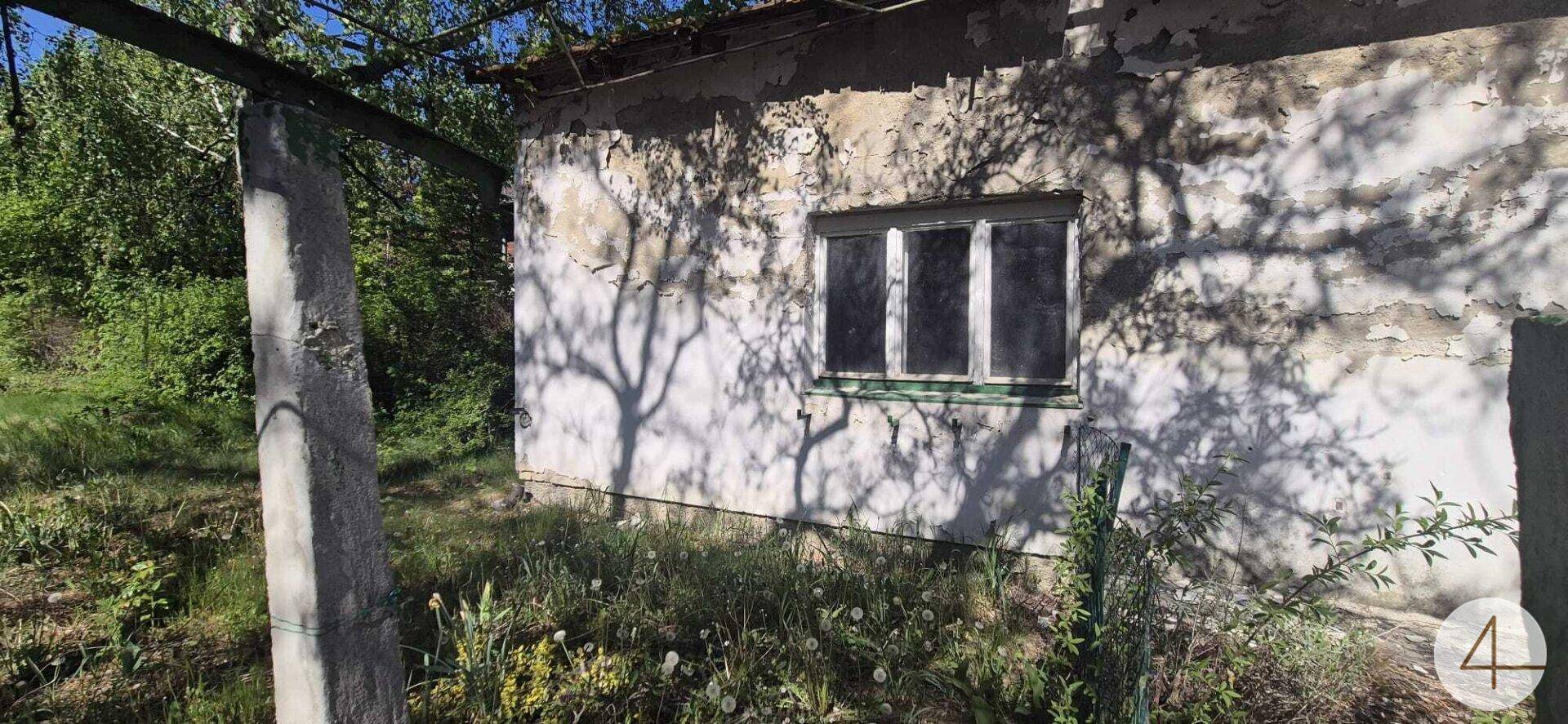 Seitenansicht eines alten Gebäudes mit abblätterndem Putz und einem Fenster, umgeben von viel Grün.