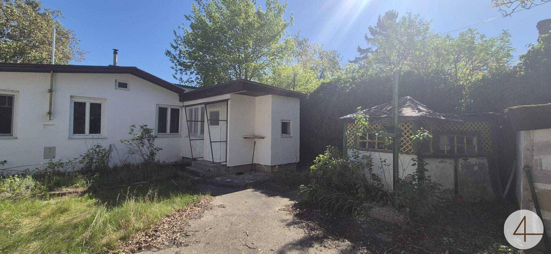Mehrere ältere Gebäude und ein verwilderter Gartenbereich unter strahlendem Sonnenschein.