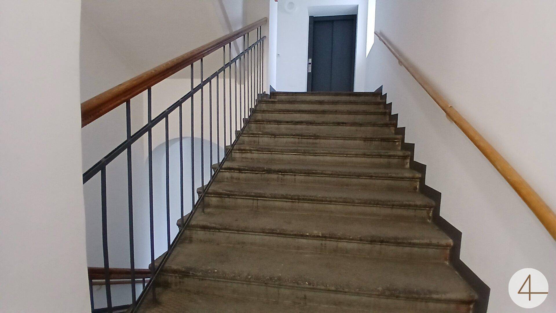 Treppenhaus mit Steinstufen und Holzgeländer, ein Aufzug ist im Hintergrund sichtbar.