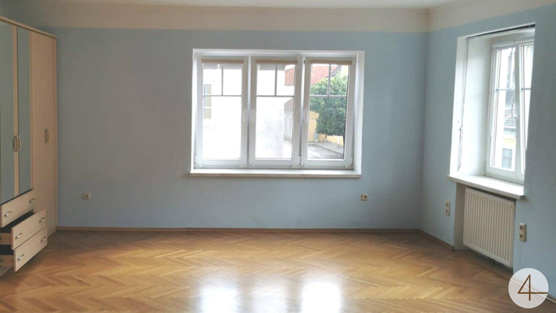 Geräumiges Zimmer mit hellblauen Wänden, Fischgrätparkett und zwei Fenstern für viel Tageslicht.
