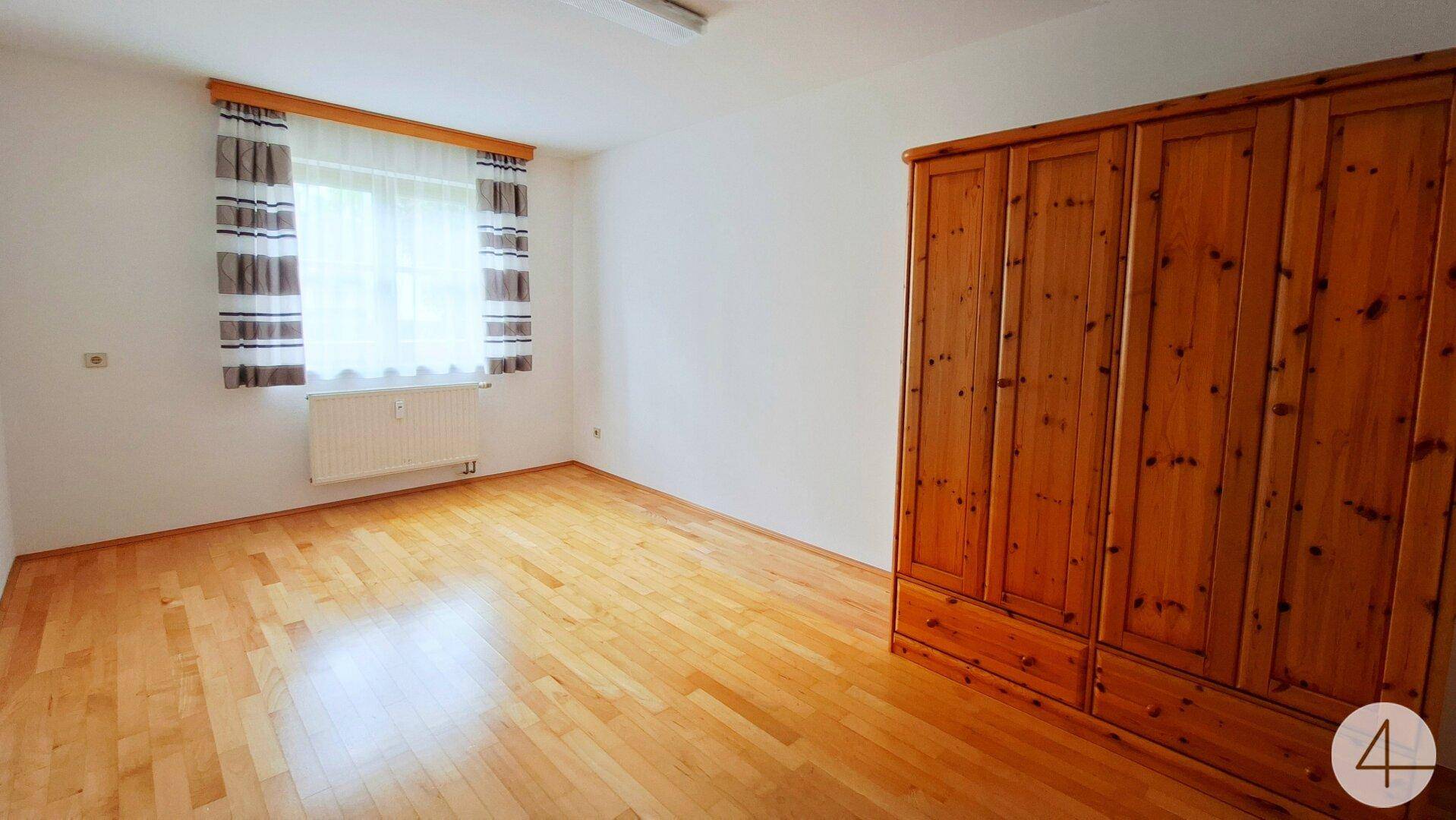 Schlafzimmer mit Holzparkettboden, Fenster und großem Holzschrank.