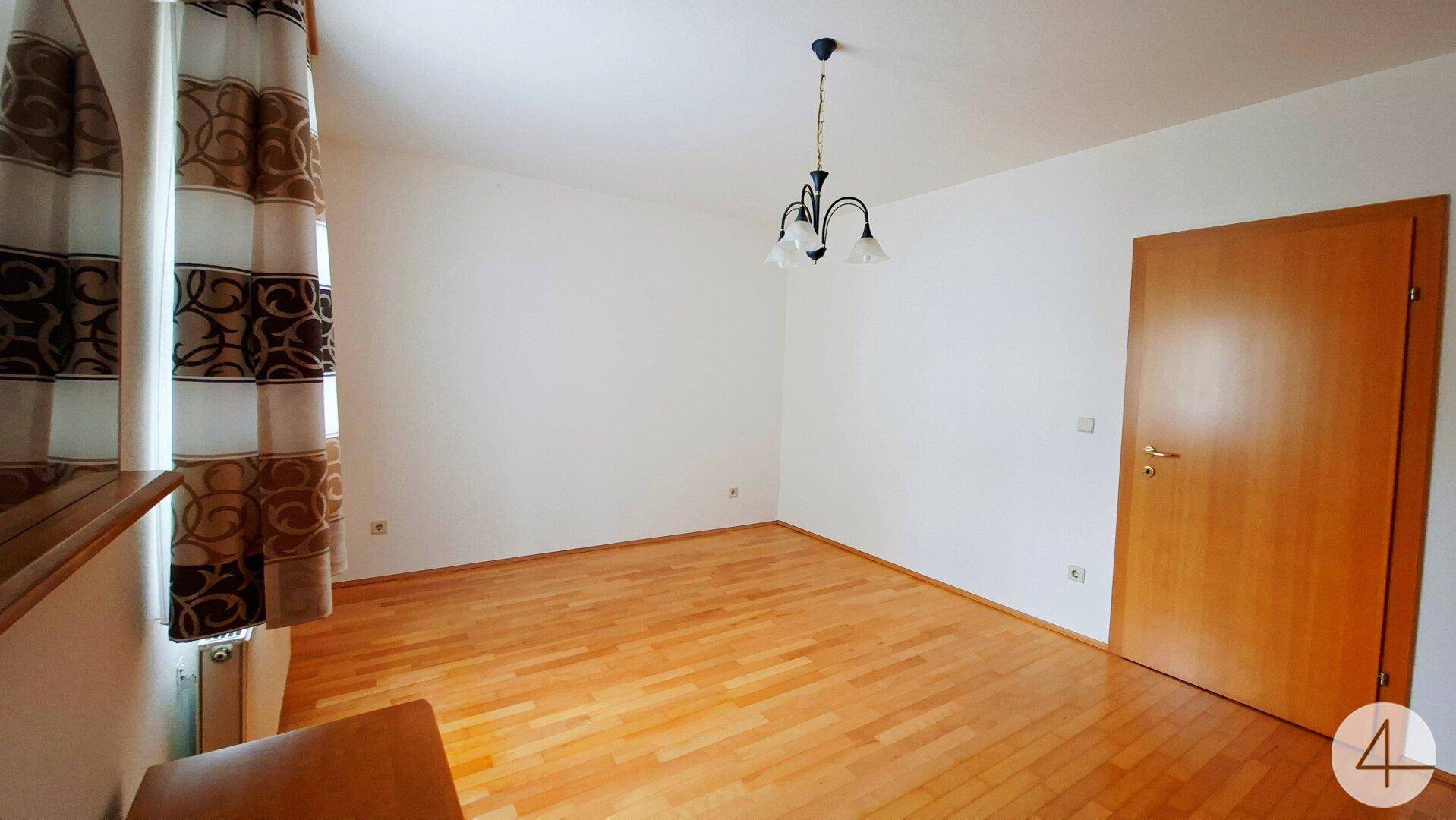 Geräumiges Zimmer mit Holzparkettboden, Fenster und Holztür.