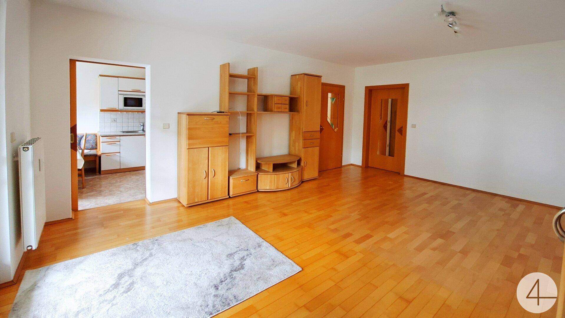 Helles Wohnzimmer mit Parkettboden und Holzmöbeln, mit Blick in eine funktionale Küche.