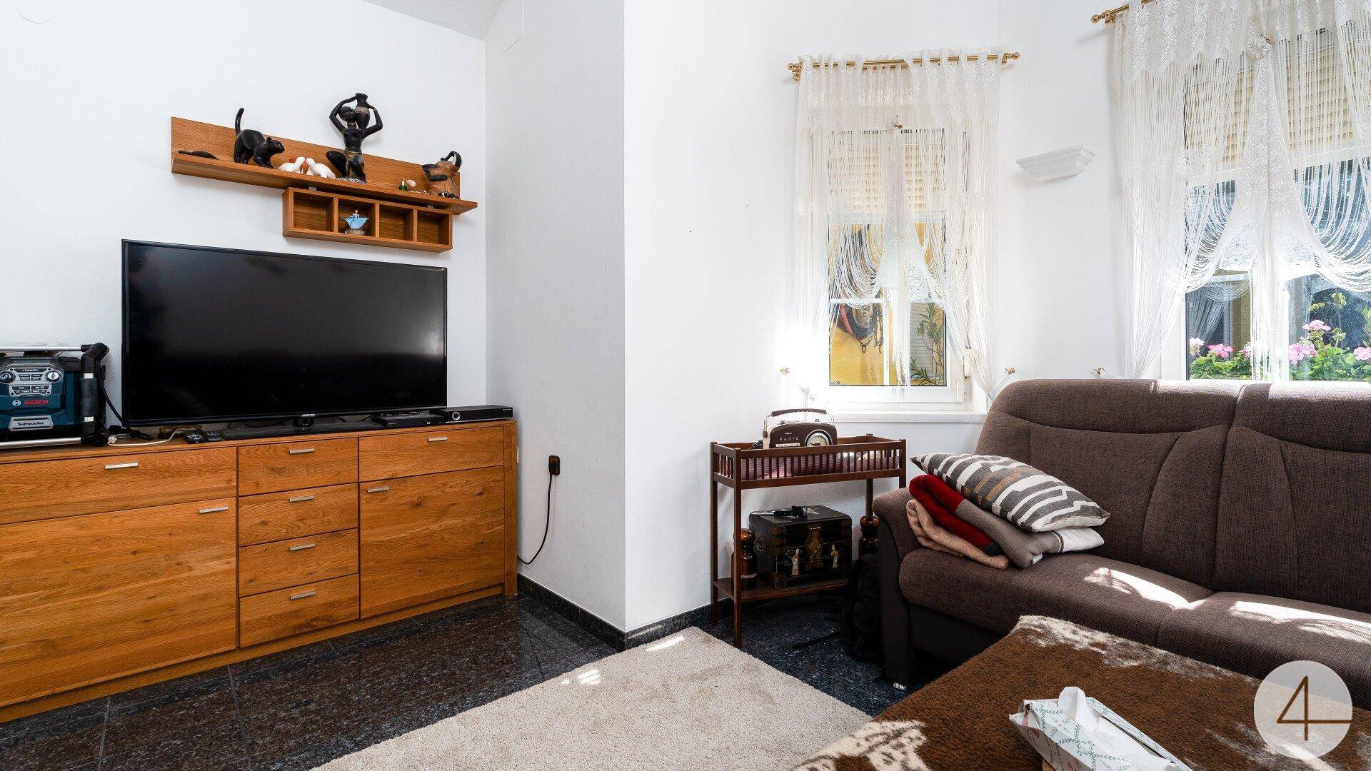 Wohnbereich mit Sofa, Fernseher auf einem Holzschrank und hellen Vorhängen am Fenster.