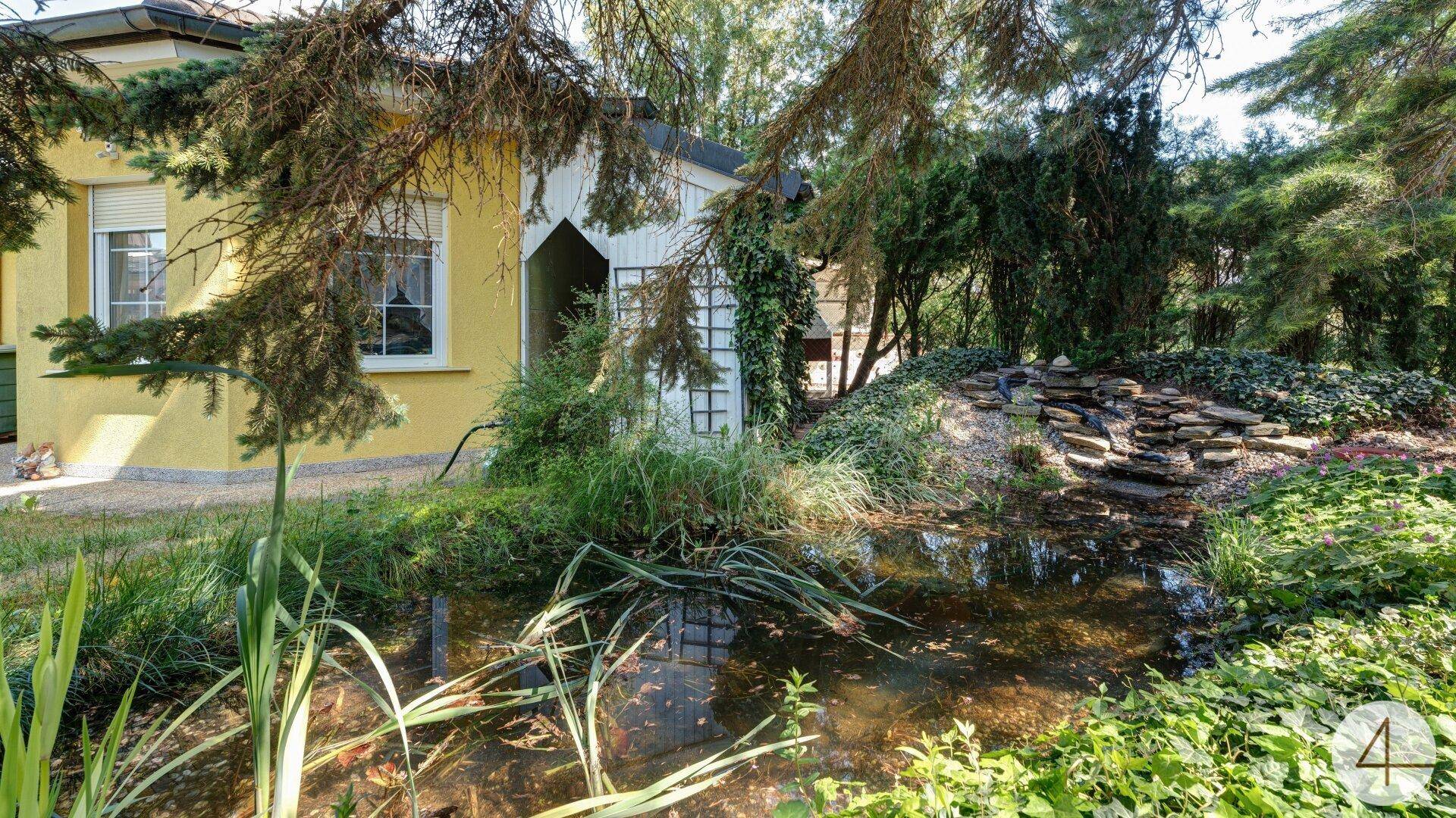 Idyllischer Gartenbereich mit einem Teich, kleinem Wasserfall und üppiger Vegetation unter hohen Bäumen.