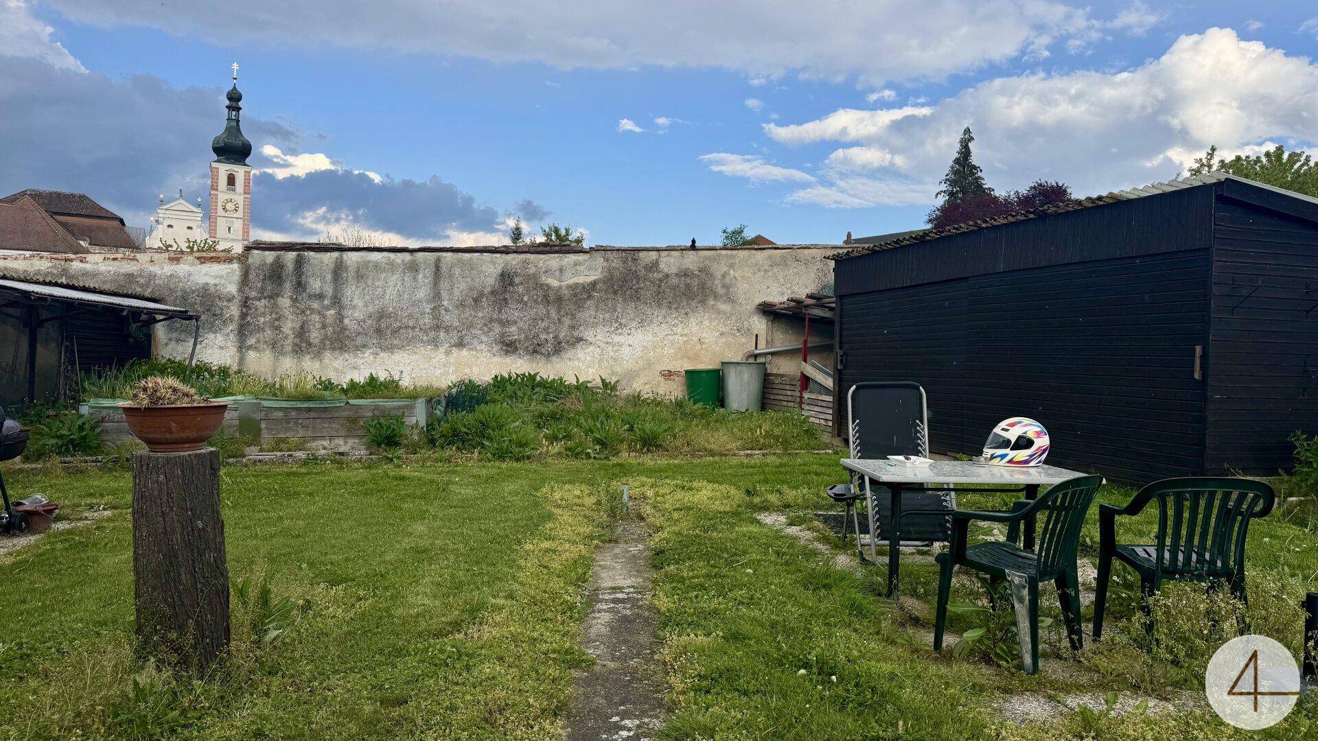 Einfacher Gartenbereich mit Sitzgelegenheiten, Schuppen und Blick auf eine Kirche.
