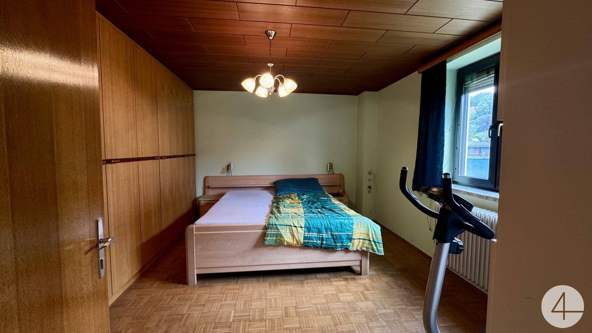 Schlafzimmer mit Holzbalkendecke, Einbauschränken und einem Heimtrainer.